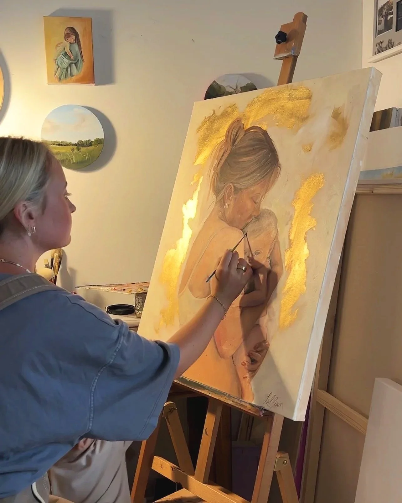 A woman painting a portrait of a mother holding a child on a large canvas in an art studio, with several landscape and portrait paintings hanging on the wall behind her.