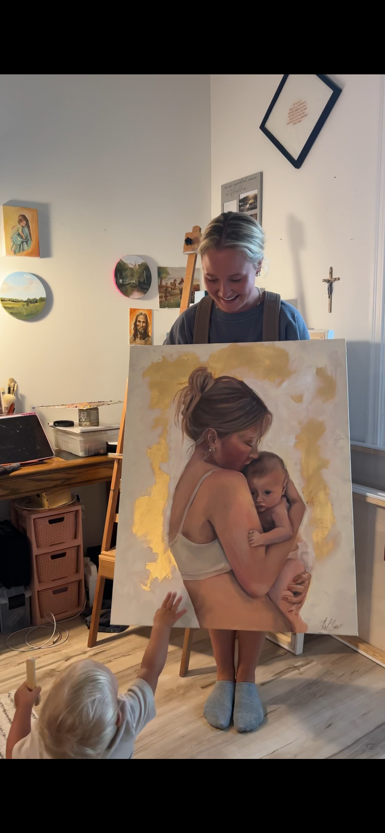 A woman is holding a realistic painting of a mother holding a baby, with a young child reaching towards it. A smiling woman stands behind the painting in a room decorated with small framed pictures and a crucifix on the wall.