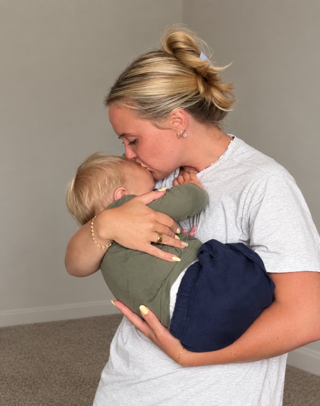 A woman with blonde hair styled in a bun kisses her young child on the forehead as she holds him close, both with light skin, in an indoor setting with a neutral gray background.