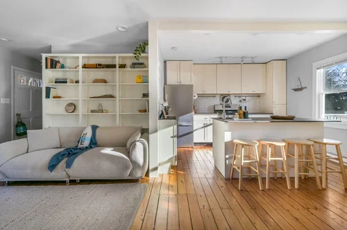 Open-concept living room and kitchen with white shelving, a beige sofa, wooden flooring, and a kitchen island with four bar stools.