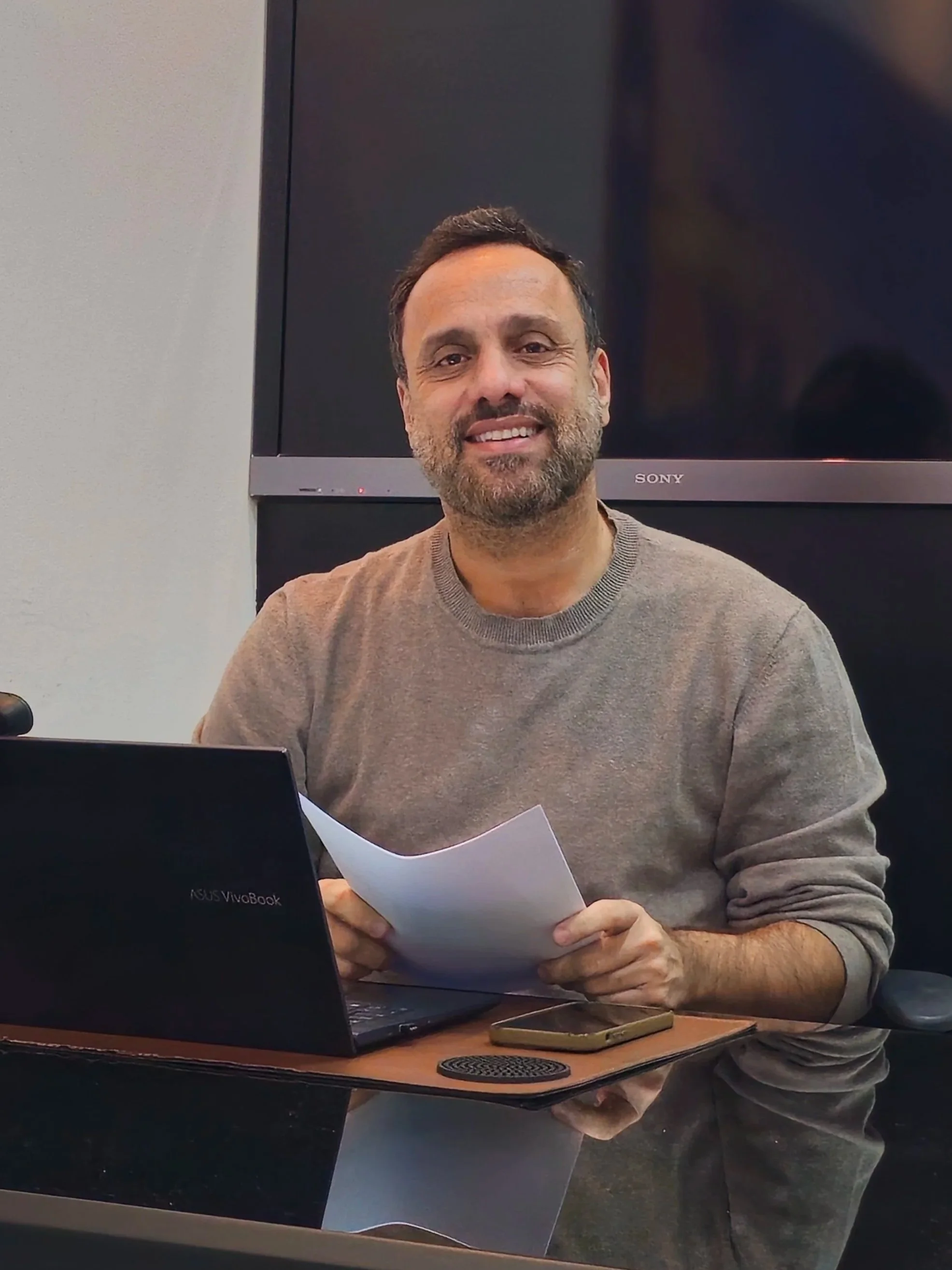 Luis Fernando Barbosa Sahium, Homem sorridente com barba, usando suéter bege, sentado em frente a um computador, segurando uma folha de papel na mão direita, ao lado de um telefone celular dourado, com uma televisão ao fundo.