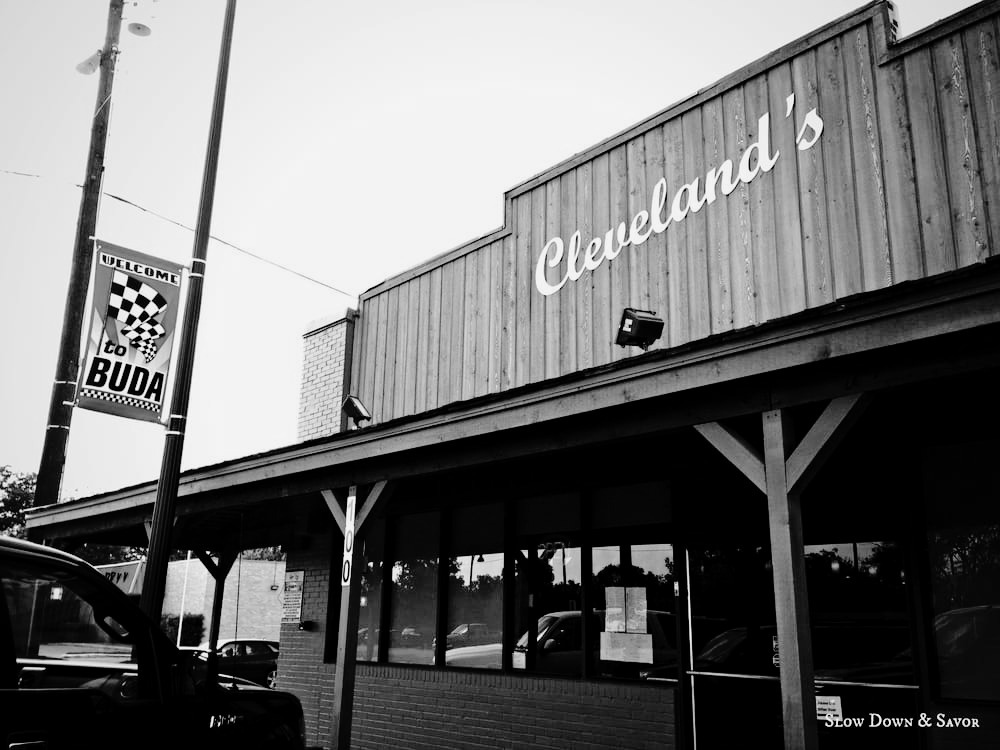 Black and white photo of a building with a large sign reading "Cleveland's" and a border with windows. A welcome sign for Buda town is on a pole next to the building. There are cars parked in front.