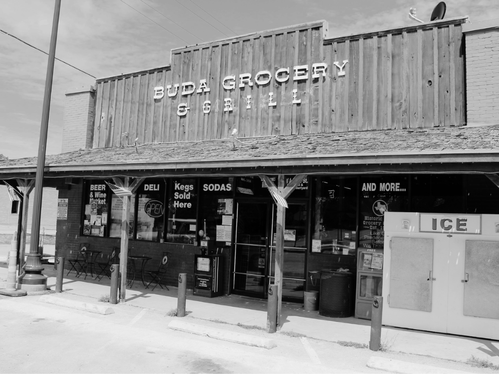 Black and white photo of Buda Grocery & Grill, a convenience store on a sunny day with outdoor seating, an ice machine, and signs for beer, wine, deli, kegs, sodas, and more.