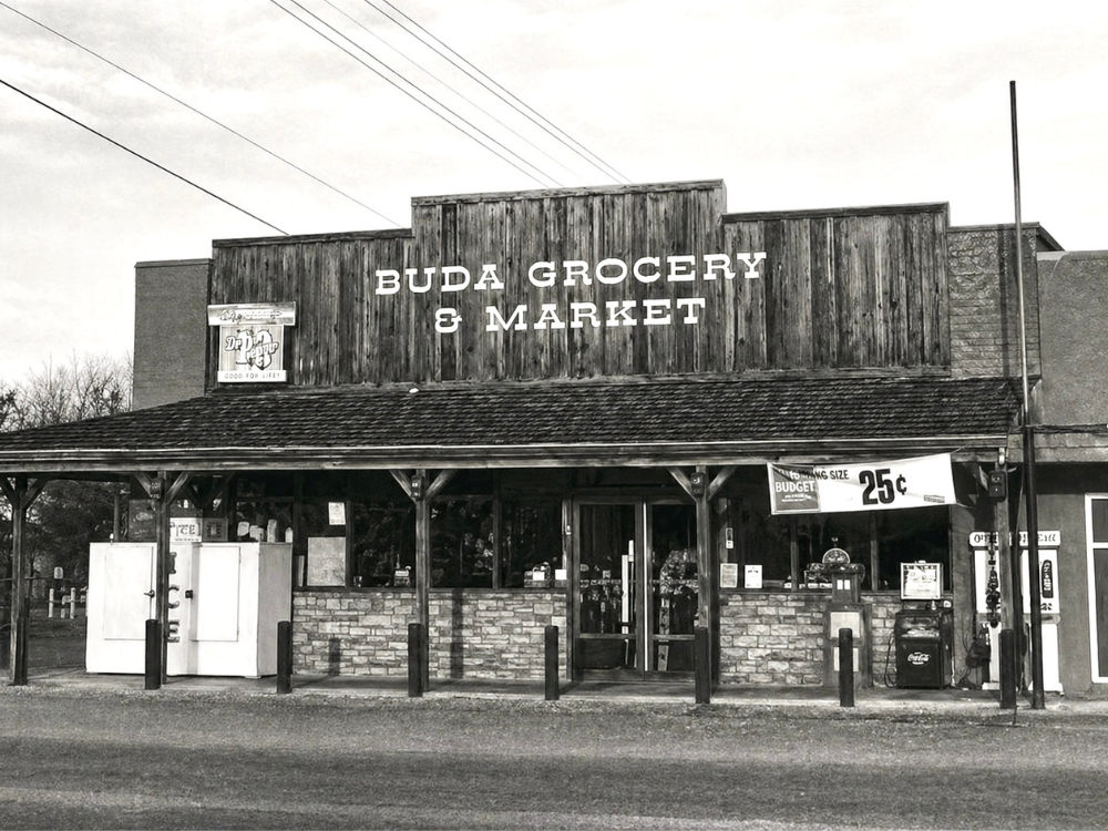 Black and white photo of Buda Grocery & Market, a small rural grocery store with a wooden facade, hanging sign, outdoor vending machines, and a sidewalk with protective posts.