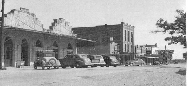 Black and white photograph of a small town street with vintage cars parked along the curb and buildings with storefronts and awnings.