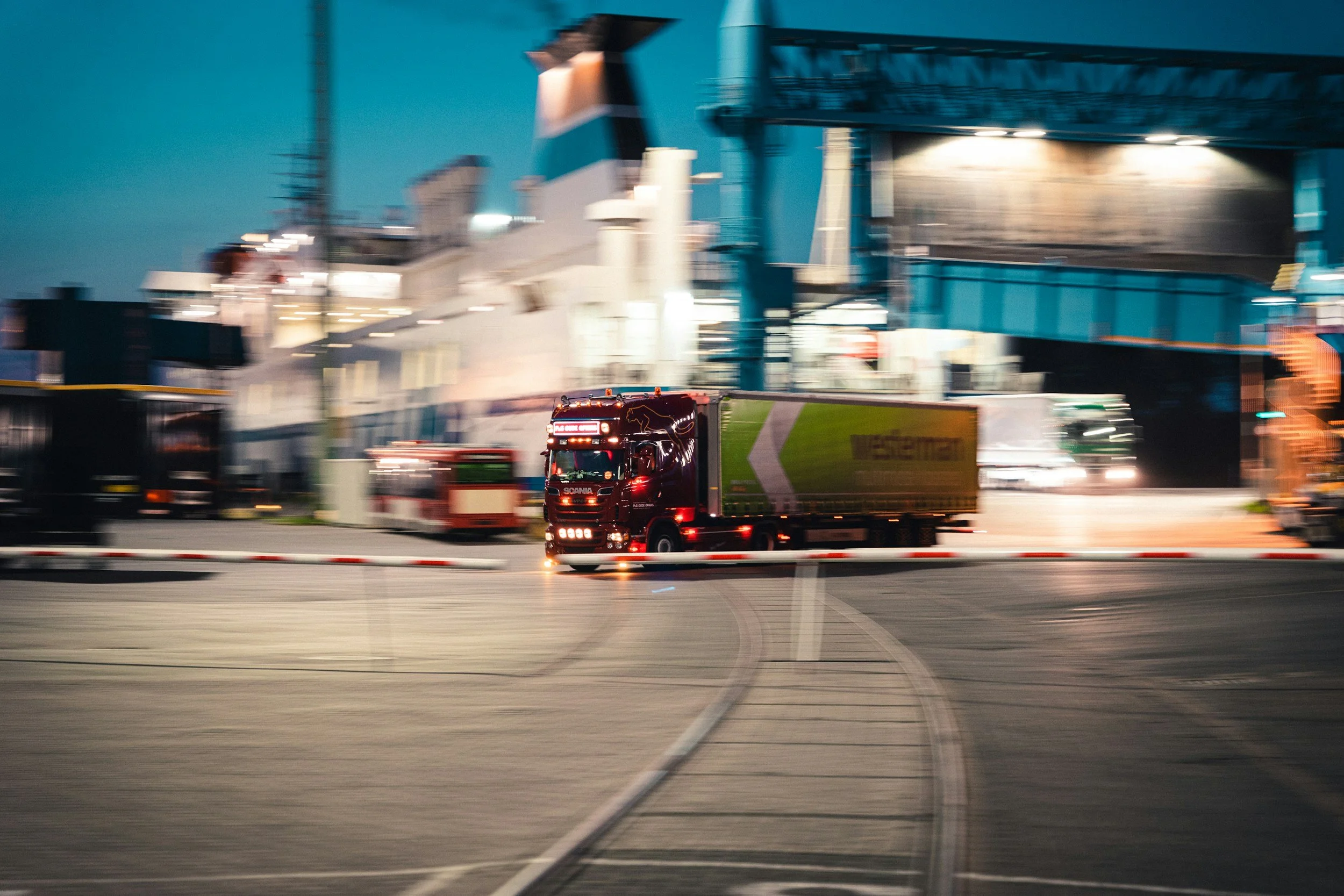 A red semi-truck driving on a highway with a blurred background of an industrial port and ships.