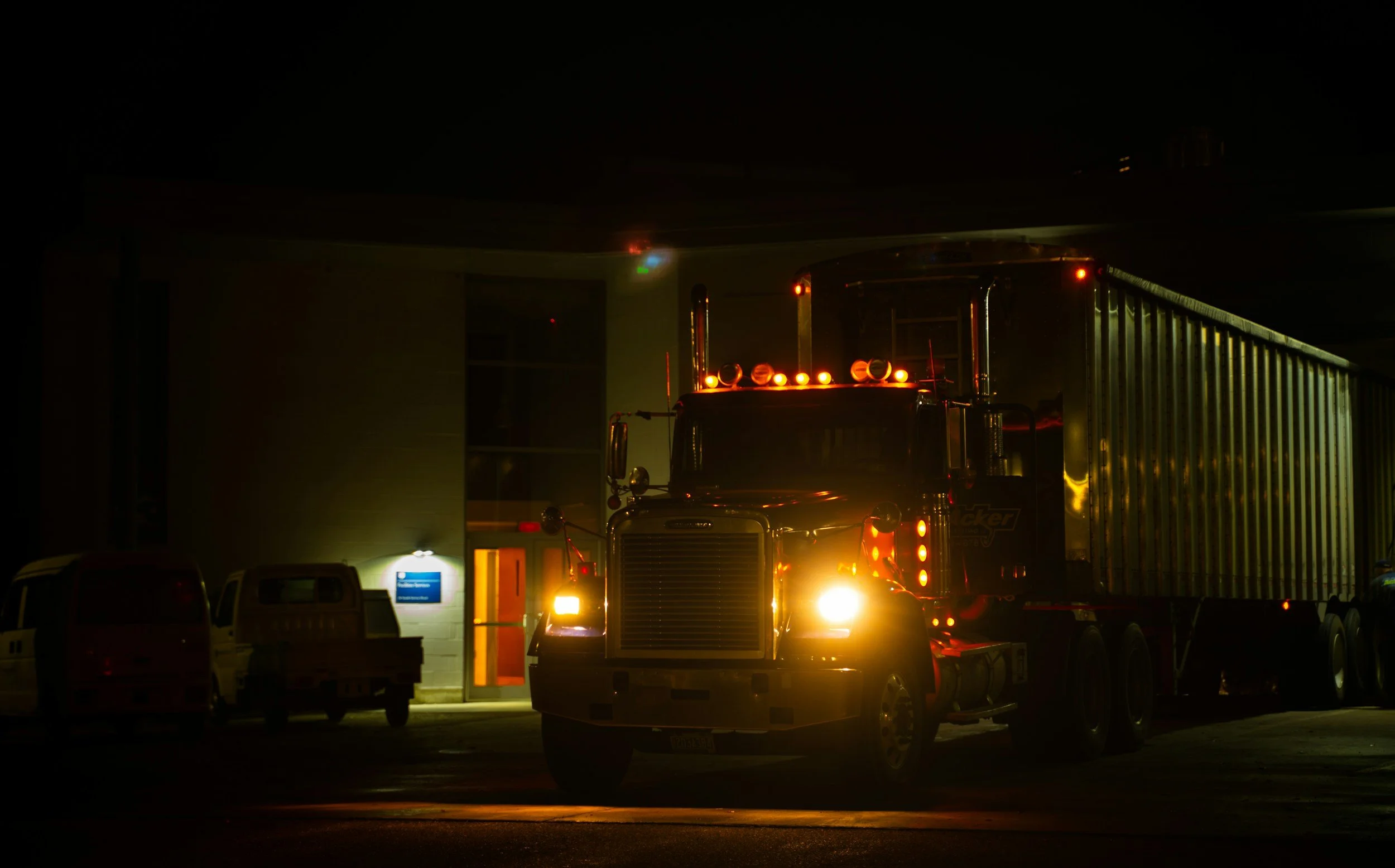 A large semi-truck with bright orange lights on the roof and front, parked outside a building at night, surrounded by other vehicles.