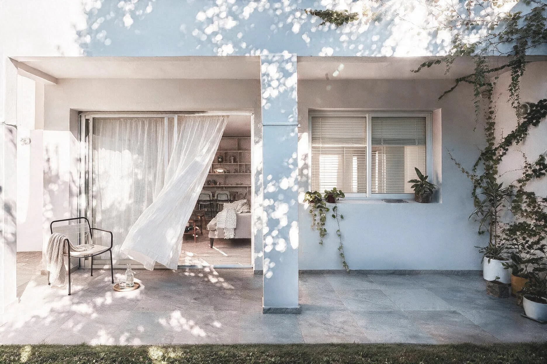 Fachada de casa blanca con balcón, jardín y puerta corrediza de vidrio que se abre a un living con plantas y sombra de árboles — reforma de vivienda en zona norte, provincia de Buenos Aires