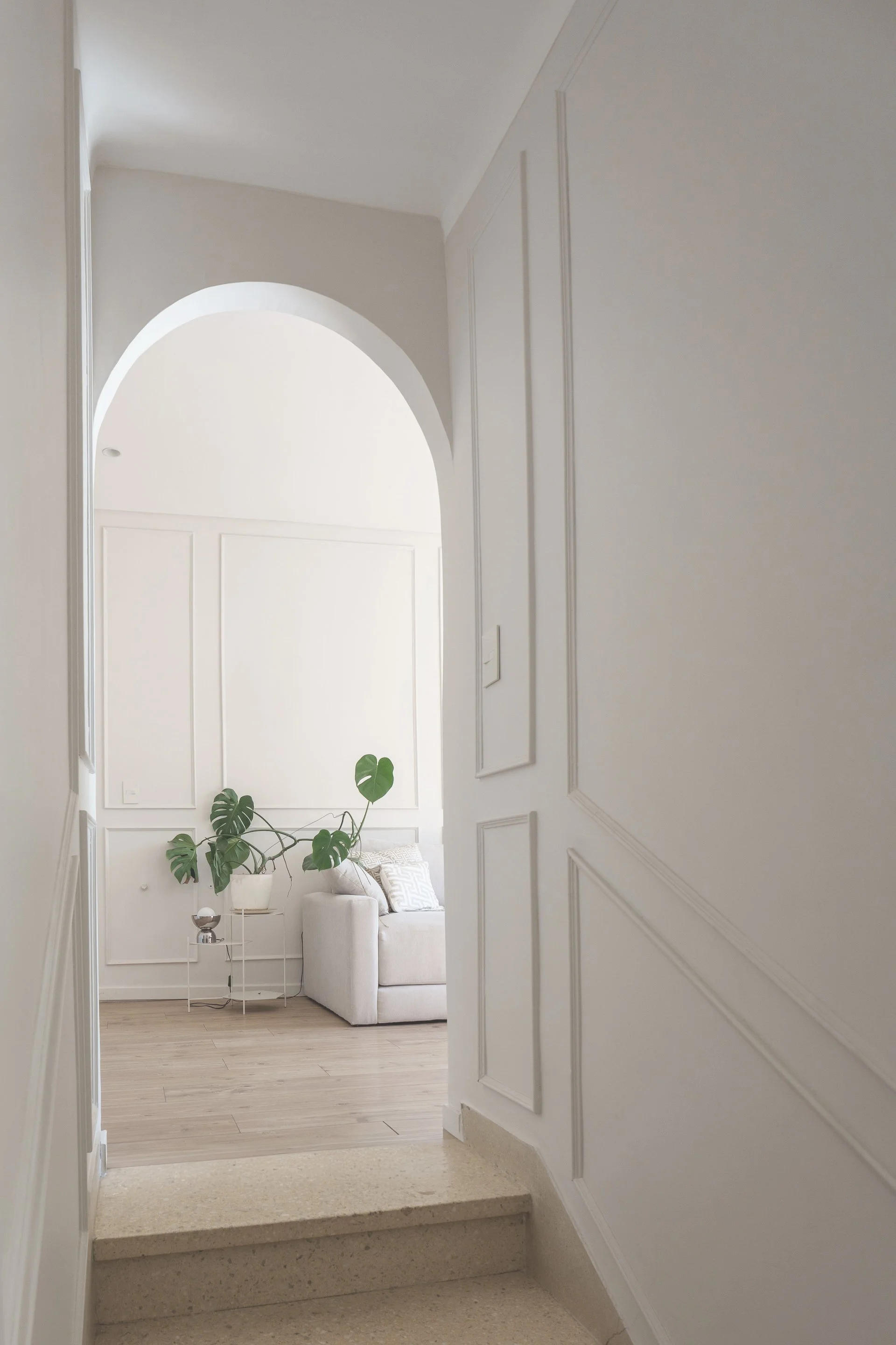 Vista hacia living luminoso con sillón blanco, planta Monstera, techos altos y piso de madera clara — proyecto de reforma en CABA