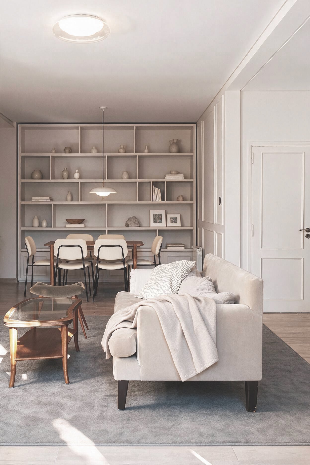 Living con sillón beige, alfombra gris y mesa auxiliar, integrado a comedor con estantería blanca y objetos decorativos — proyecto de reforma en CABA