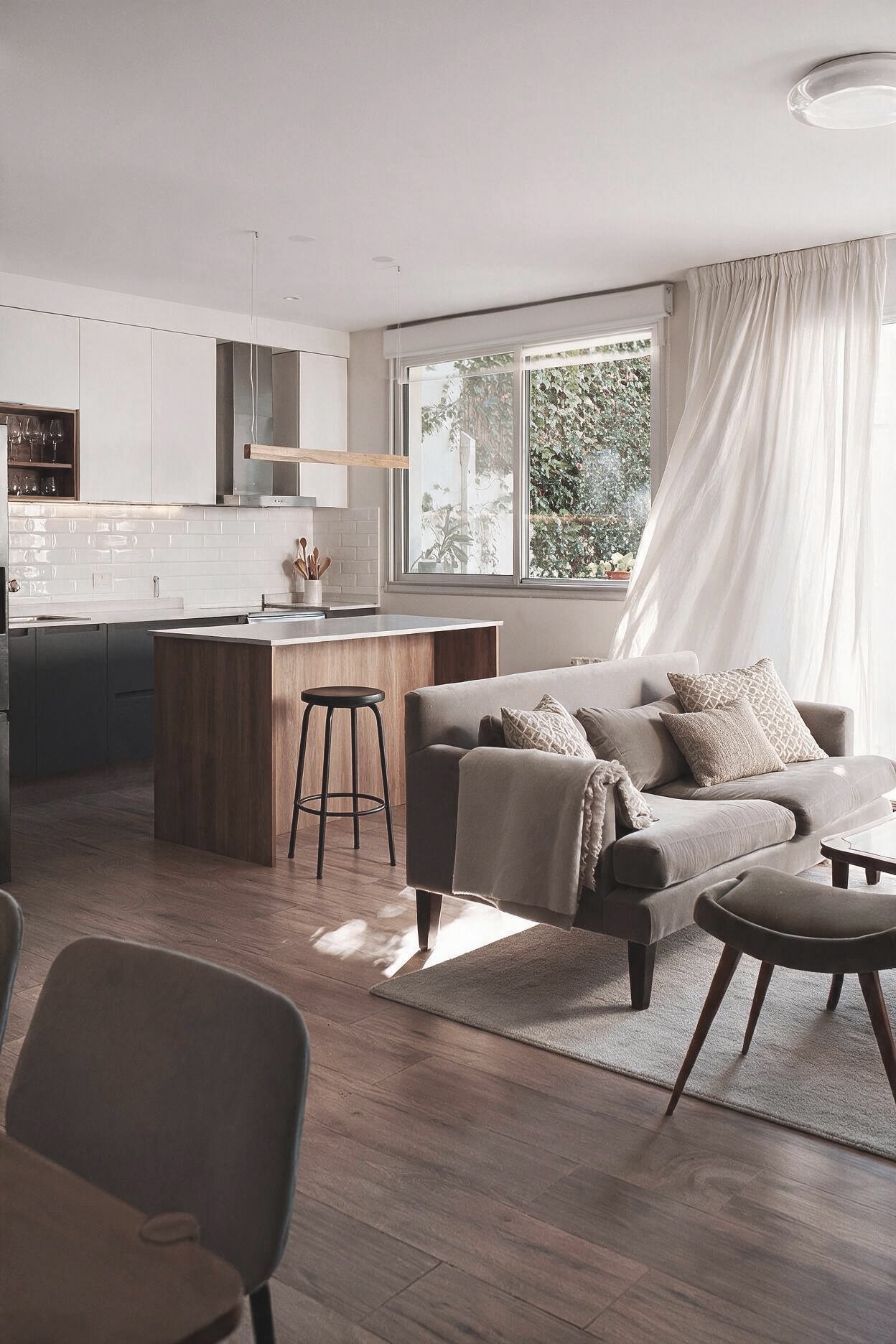 Living con sillón blanco, almohadones y manta, alfombra beige y ventanales, integrado a cocina con isla blanca y vista a jardín — proyecto de reforma en Buenos Aires