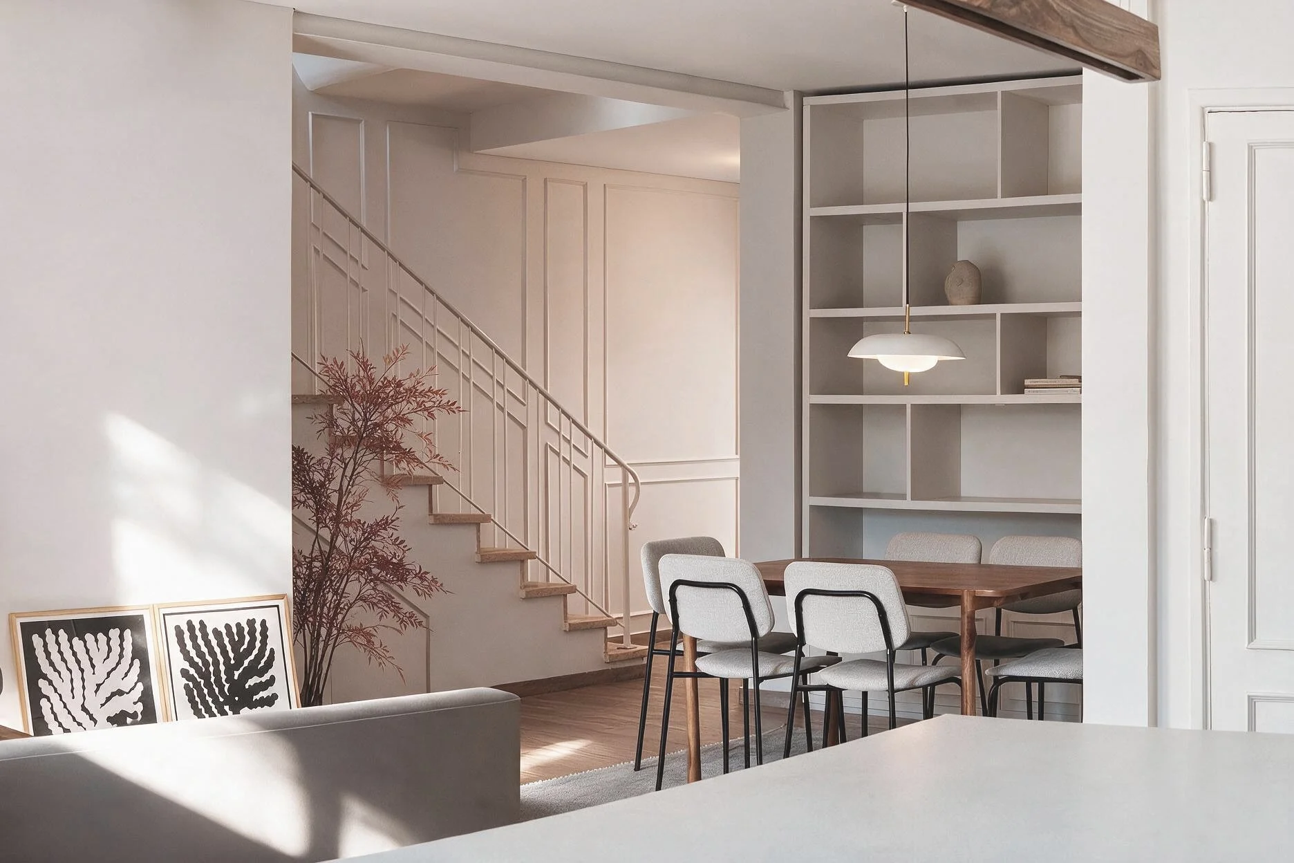 Comedor con mesa de madera, seis sillas tapizadas gris claro, estantería empotrada blanca, lámpara colgante y escalera con baranda blanca de fondo — proyecto de reforma en provincia de Buenos Aires