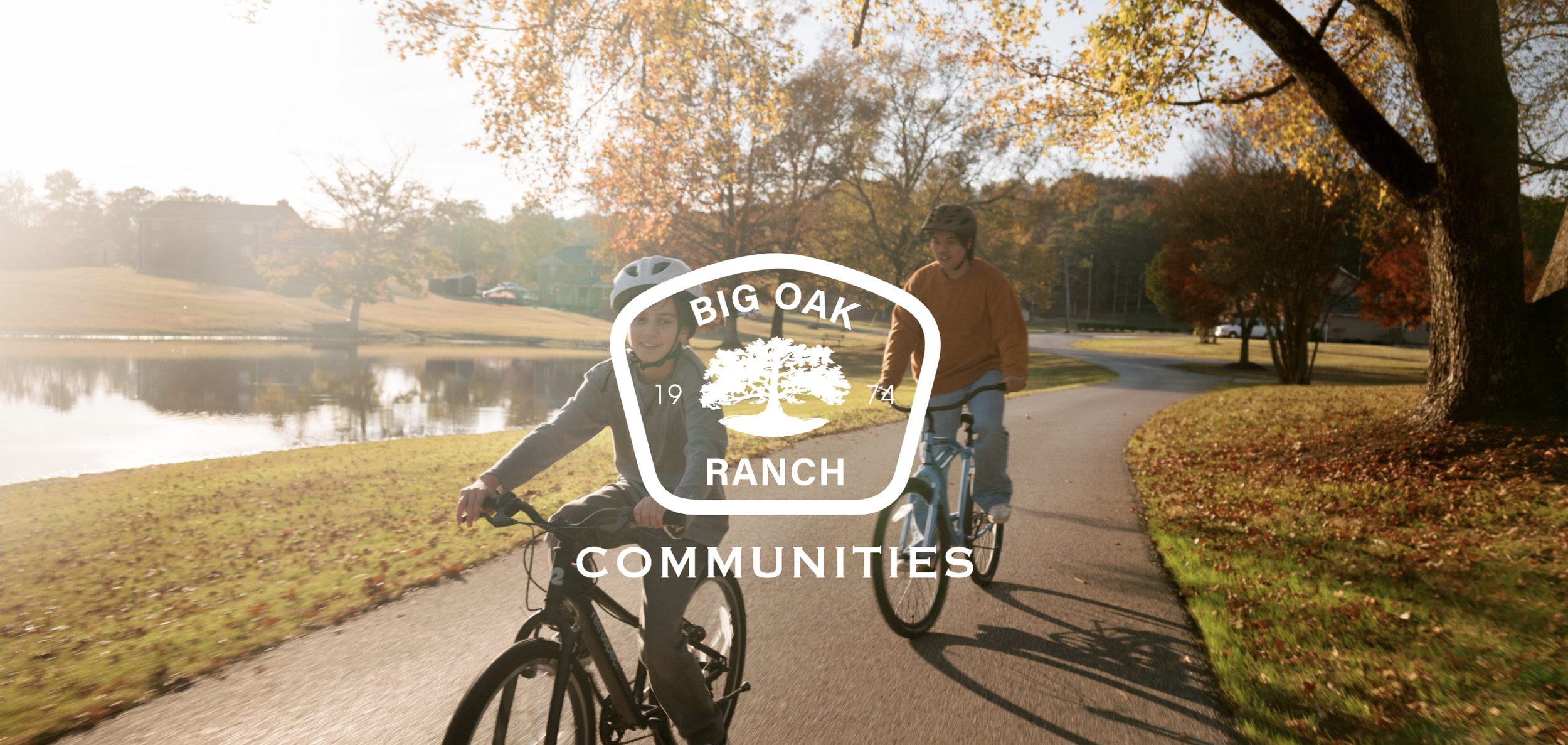 Two children riding bicycles on a scenic park trail during autumn, with colorful fall leaves on trees and a pond nearby, overlaid with the Big Oak Ranch Communities logo.
