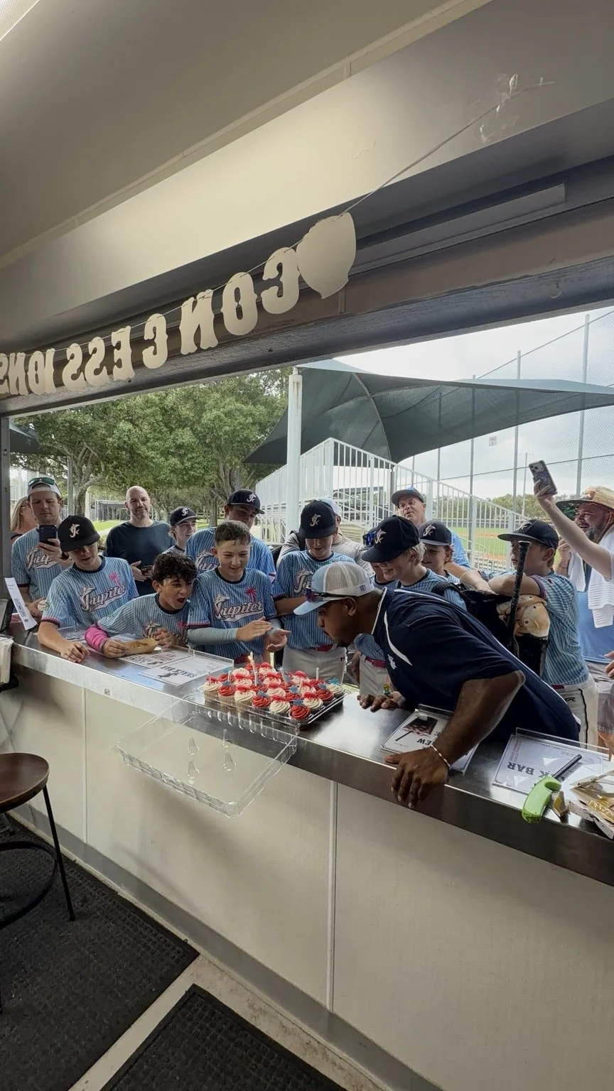 Coach Renny Hidalgo blowing out birthday candles surrounded by Jupiter Hammerheads players at the concession stand.