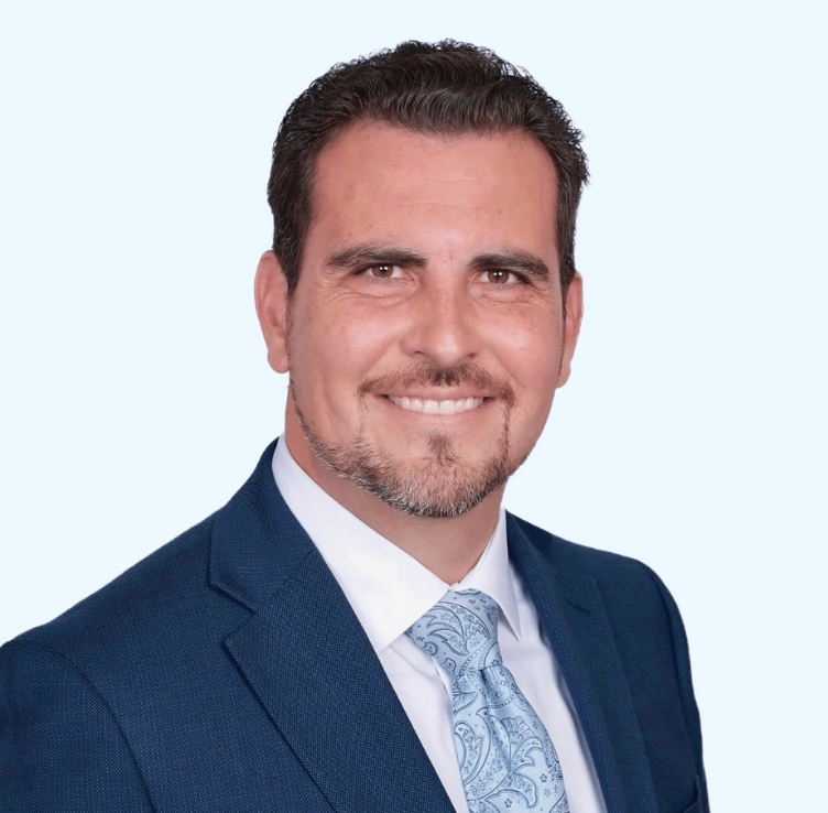 Portrait of a man in a navy suit, white shirt, and patterned light blue tie, smiling against a light background. This portrait is of Ashley Martinez, Founder and Principal of CPRA Strategies.