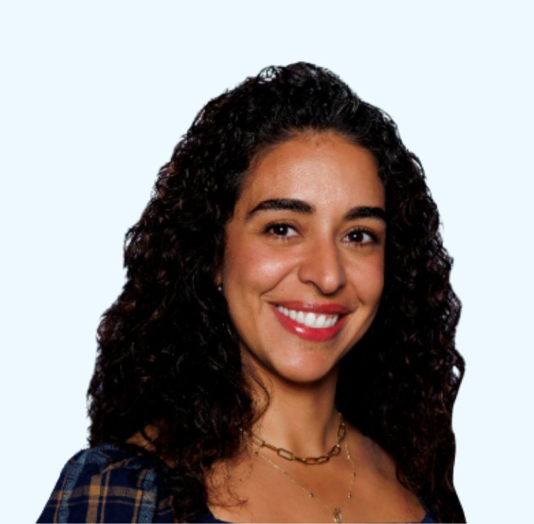 Portrait of a woman with curly dark hair smiling, wearing a checkered shirt and layered necklaces, against a light background. A woman with curly dark hair smiling, wearing a necklace and a plaid shirt, against a light background. This portrait is of