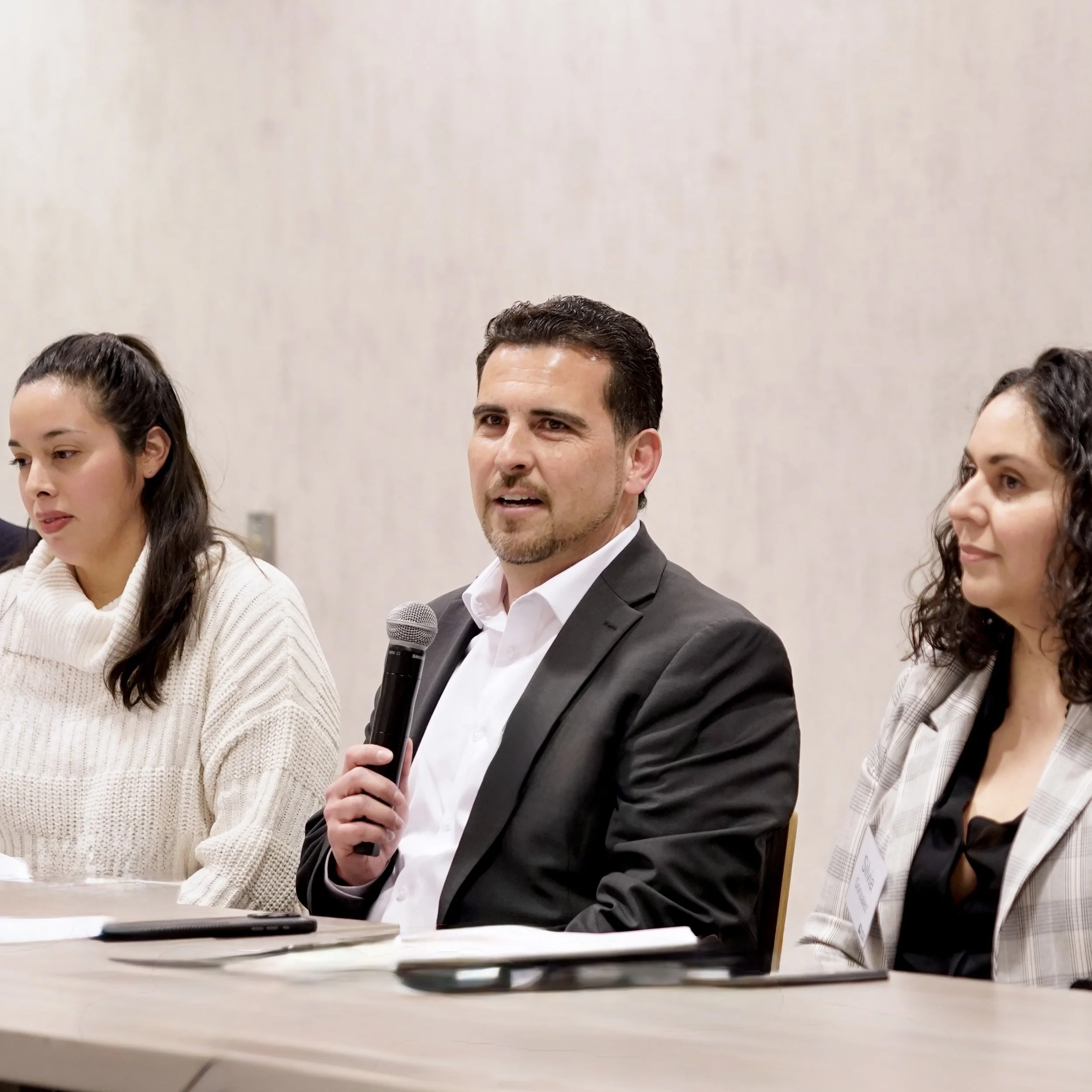A group of five people sitting at a table during a formal event or meeting. The central figure is a man named Ashley Martinez who has dark hair and a beard, wearing a dark suit and white shirt, holding a microphone.