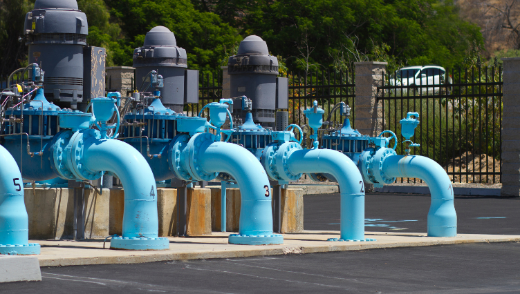 A row of large blue industrial water pipes with numbered labels, connected to pumps, in an outdoor facility with a black asphalt ground and a fenced area with trees and a car in the background.