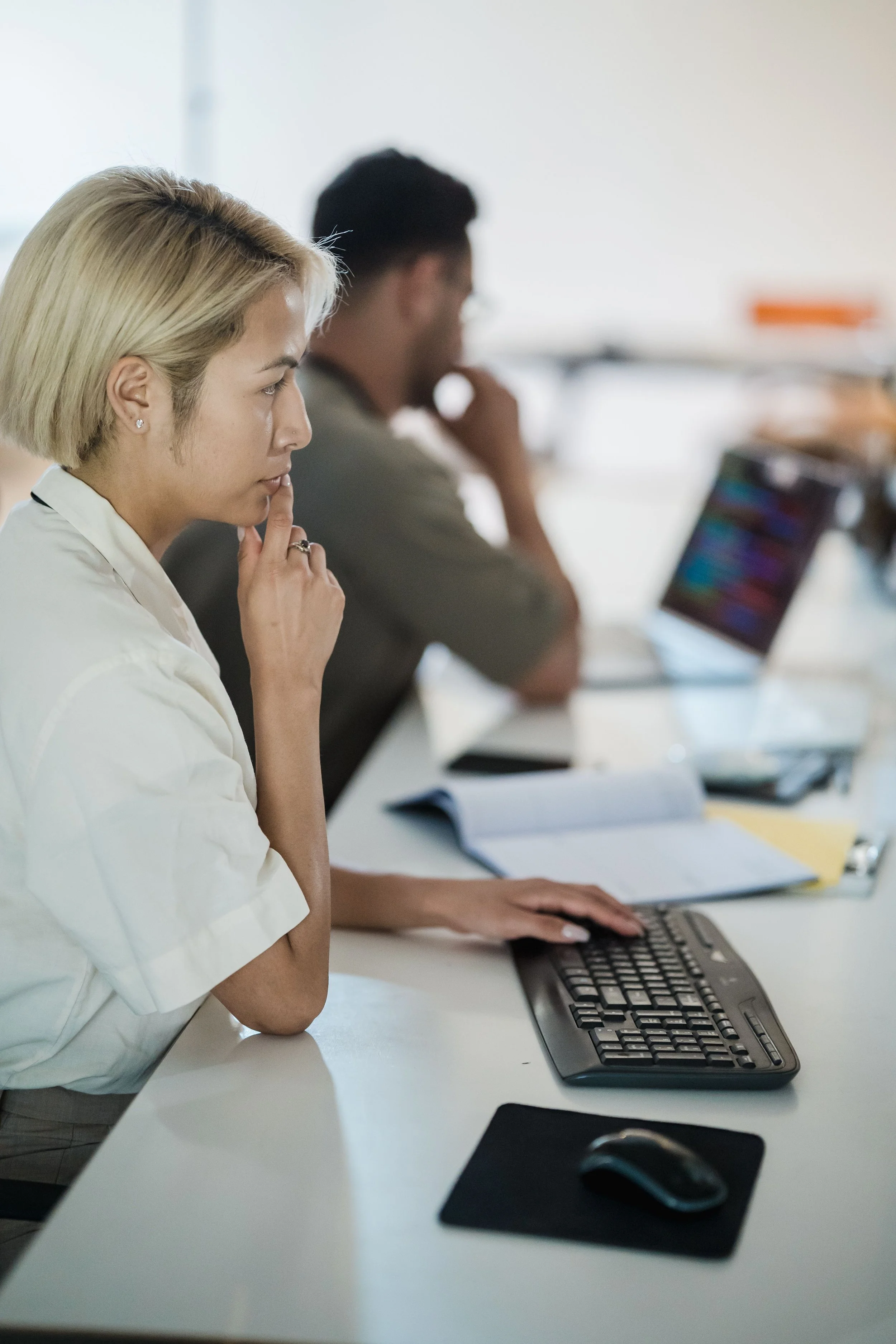 A woman with blonde hair in a white shirt sitting at a desk, deep in thought, with her hand touching her face. Behind her is a man working at a computer. The setting appears to be an office or classroom with a keyboard, mouse, and notebooks on the desk.
