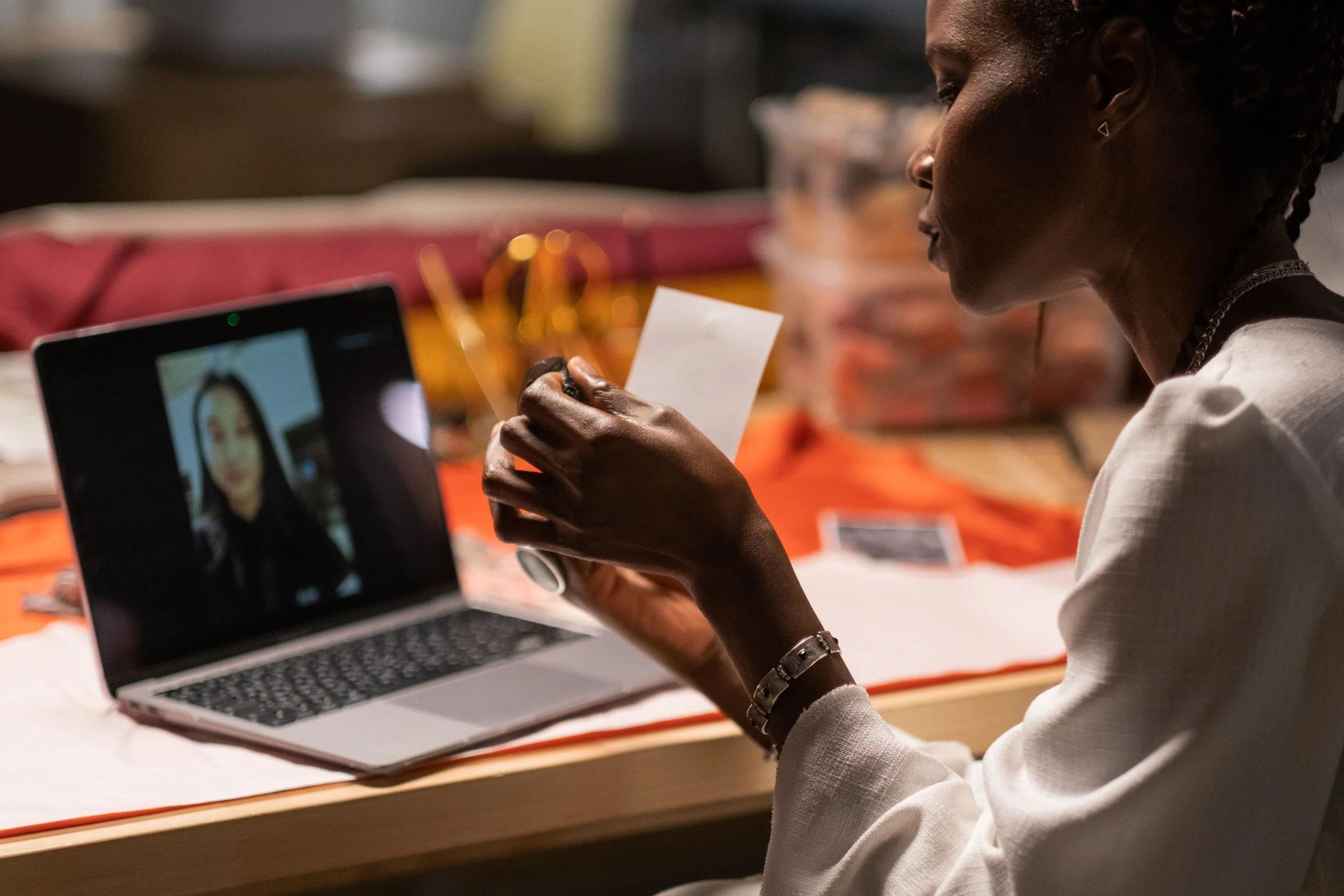 A woman with curly hair and earrings holds a small card while participating in a virtual meeting on a laptop. The screen shows another woman's face.