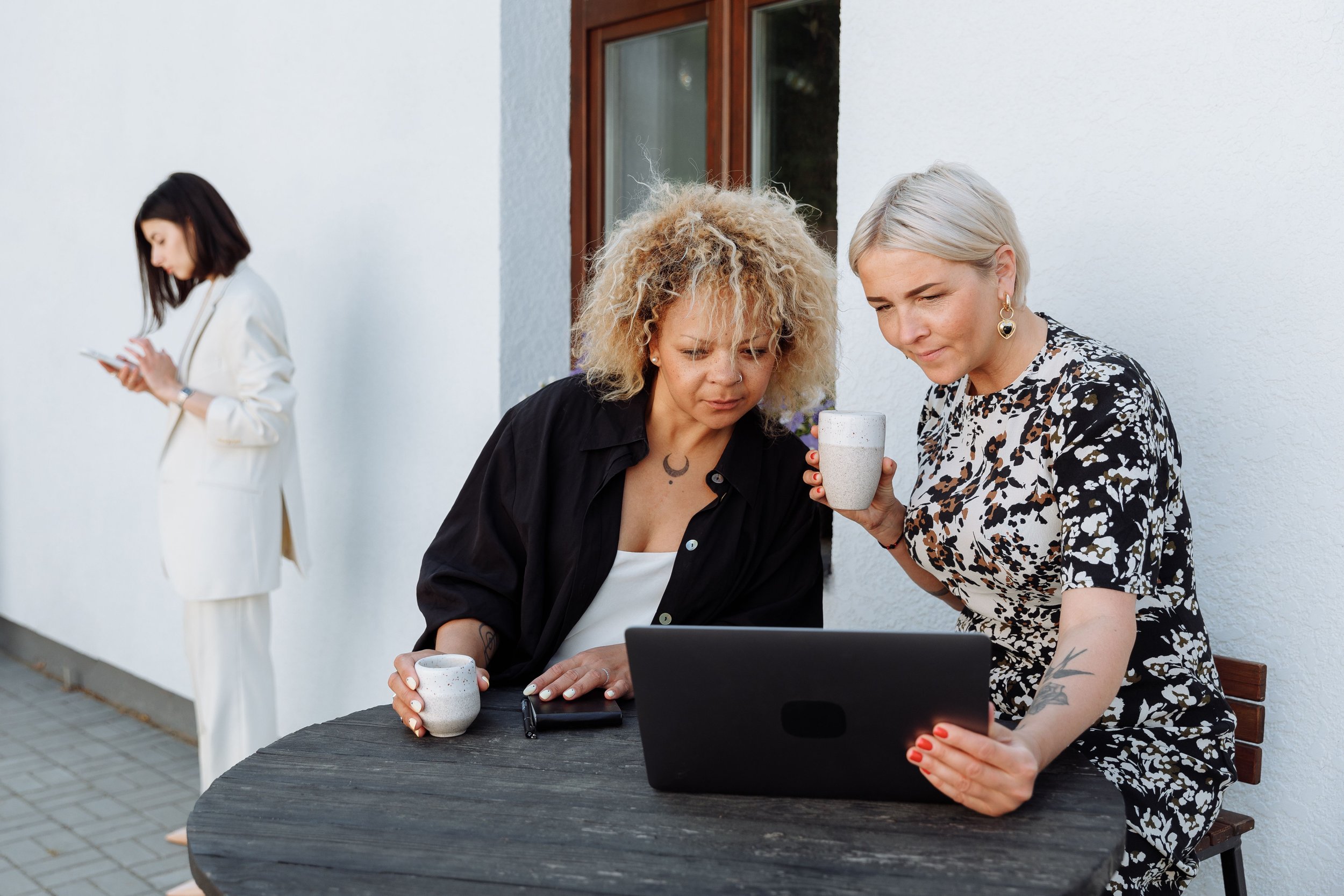 Three women are gathered outside next to a white wall, two of them are looking at a laptop on the table, while a third woman in the background is standing and using her phone.