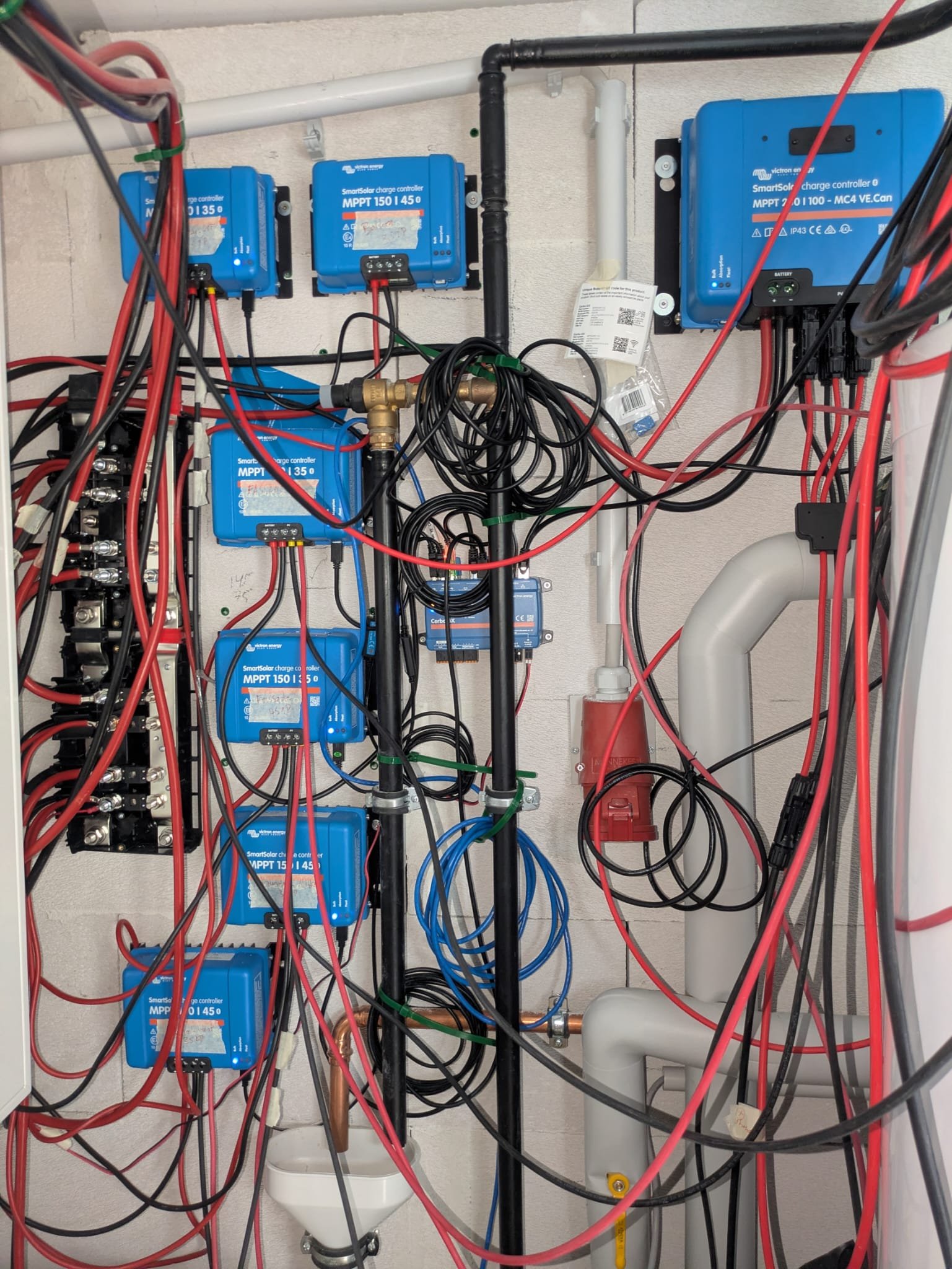Eine Wand mit mehreren blauen Solar-Ladereglern, schwarzen und roten Kabeln, einem Wasserrohr und anderen elektrischen Komponenten. Der Raum scheint für die elektrische Verkabelung und Steuerung einer Solaranlage oder ähnlichen Technologie eingerichtet zu sein.