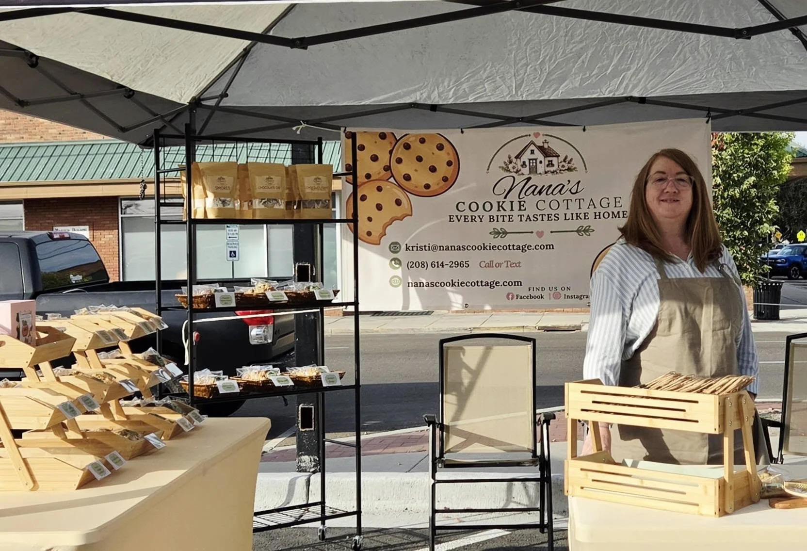 A woman standing behind a wooden display table at an outdoor market stall for Nana's Cookie Cottage, with shelves filled with cookie packs and a large banner featuring cookies and contact information in the background.