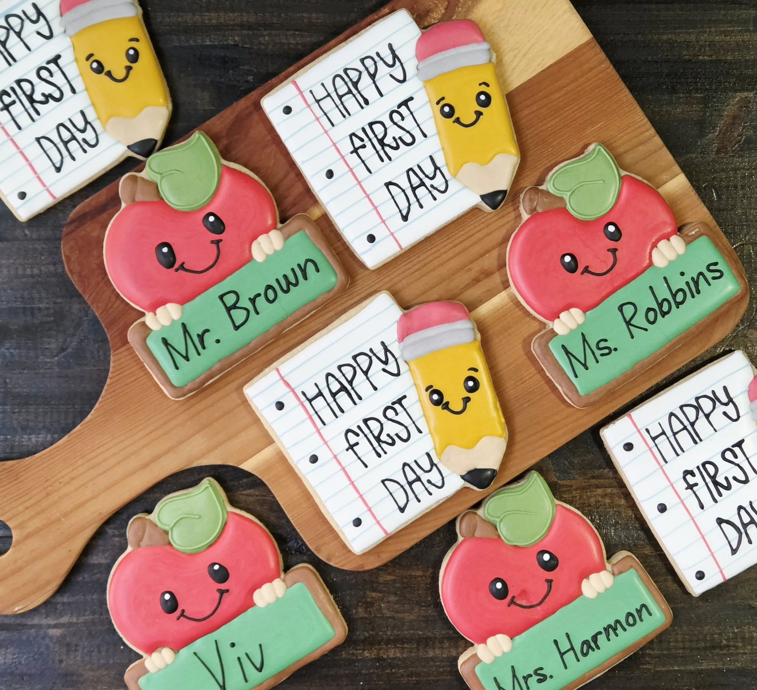 Decorated cookies with cheerful faces, holding signs with words like "Mrs. Robbins" and "Mr. Brown" on a wooden tray. Some cookies are shaped like apples, and others resemble yellow pencils. One cookie says "Happy First Day" written in black icing on