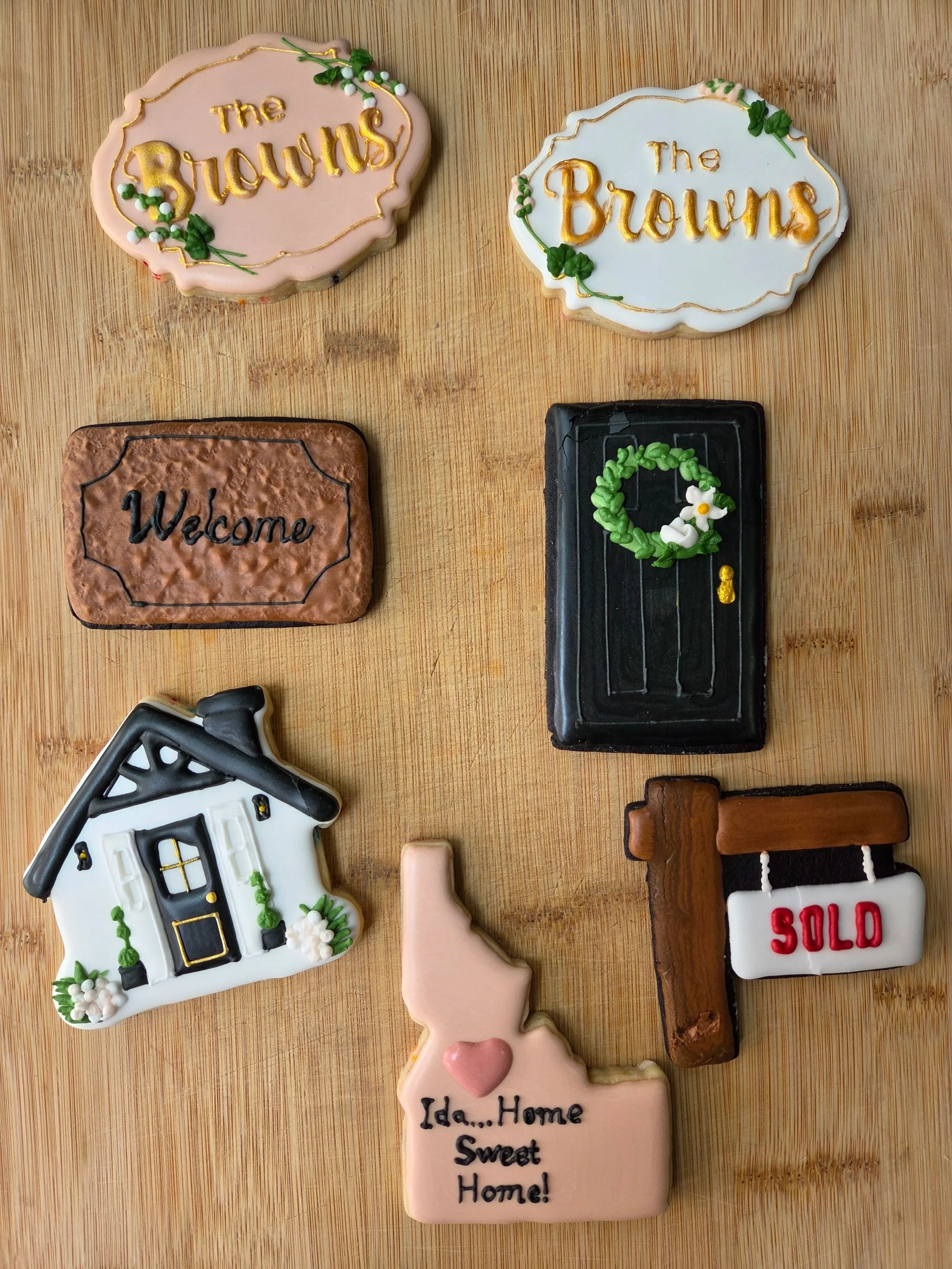 Decorative cookies shaped like a house, a sign reading "The Browns," a sign reading "Welcome," a sign reading "Sold," a pink animal-shaped cookie, and a sign saying "Ida...Home Sweet Home!" on a wooden surface.