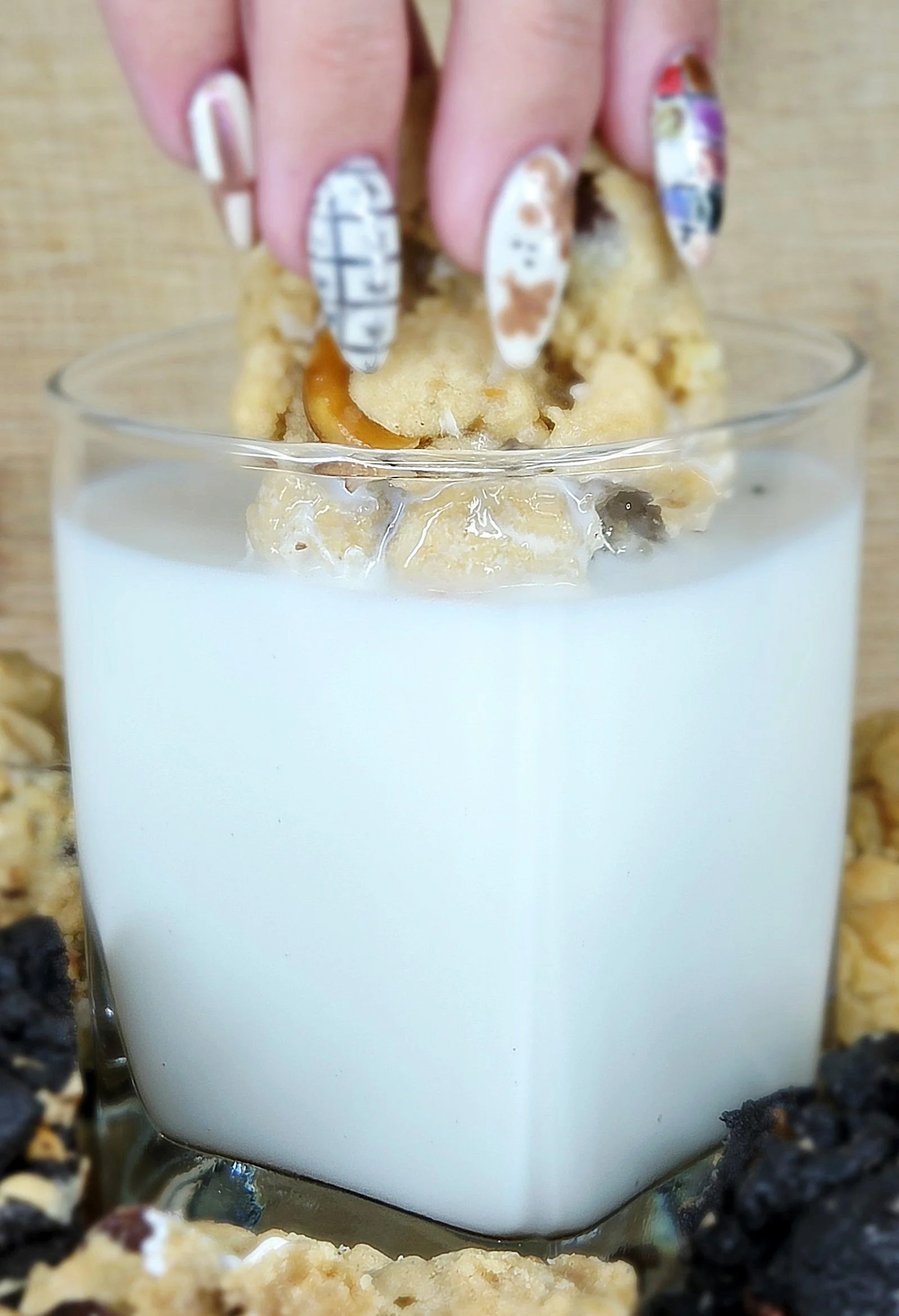 Close-up of a hand with decorated fingernails pressing onto a cookie atop a glass of milk, with cookies and berries around the glass.