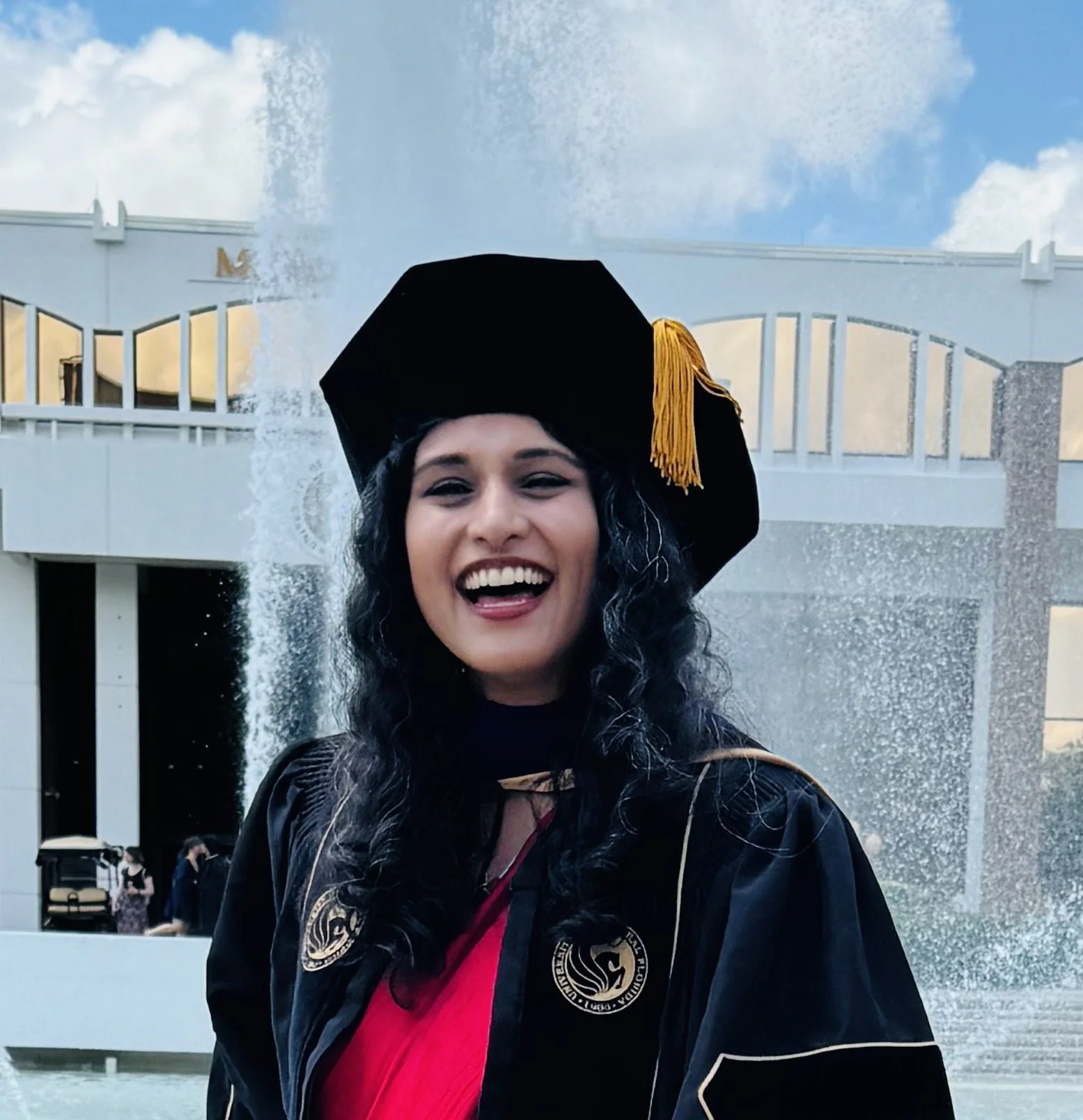 Smiling graduate in black doctoral regalia and tam with a gold tassel standing in front of a campus fountain