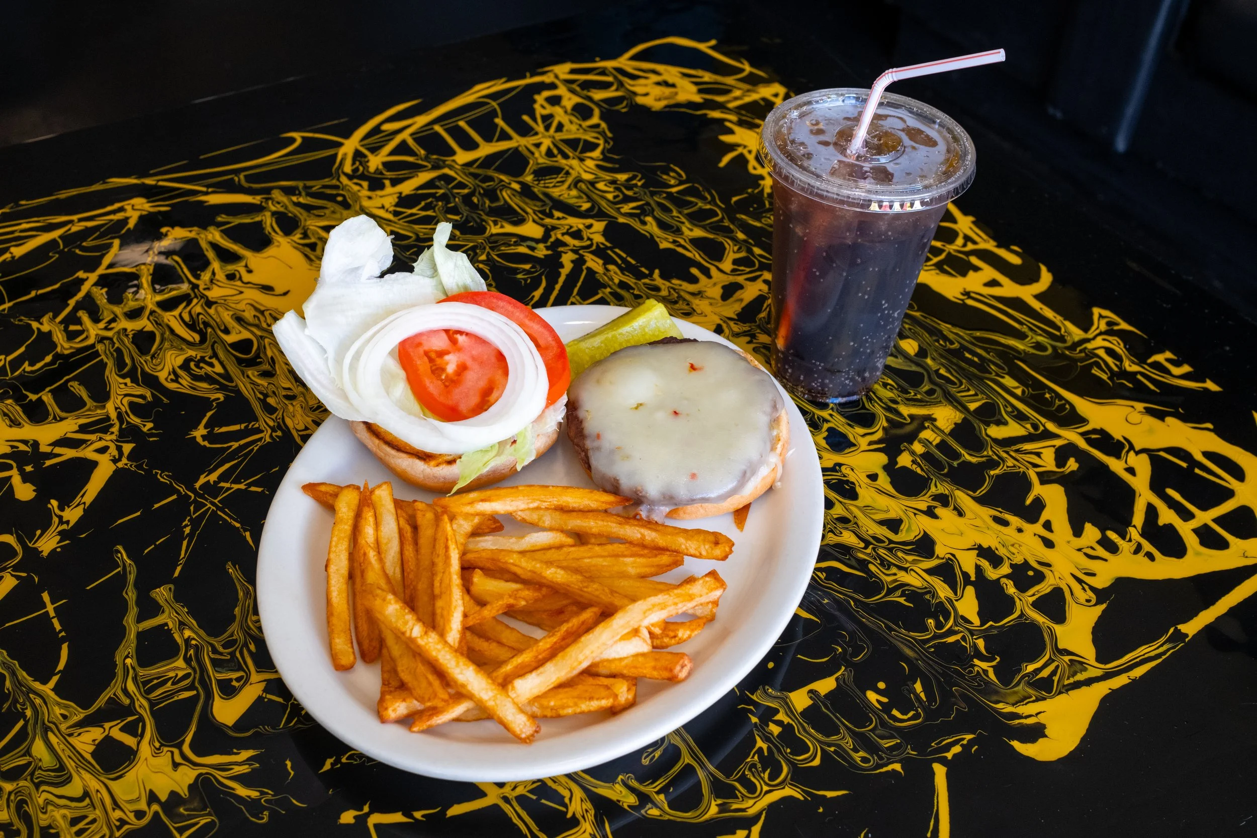 A plate of food with French fries, a burger with cheese, lettuce, tomato, and onion, and pickles. There is also a soft drink in a plastic cup with a straw next to the plate. The background features a black table with a yellow abstract pattern.