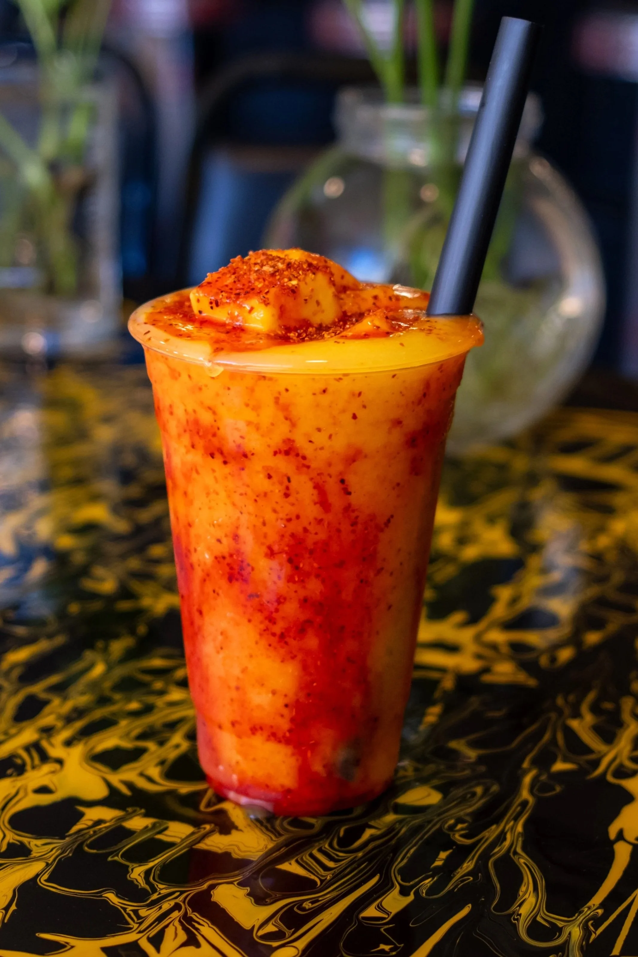 Mangonada. A colorful fruit smoothie in a clear cup with a black straw, placed on a black and yellow marbled surface, with plants in the blurred background.