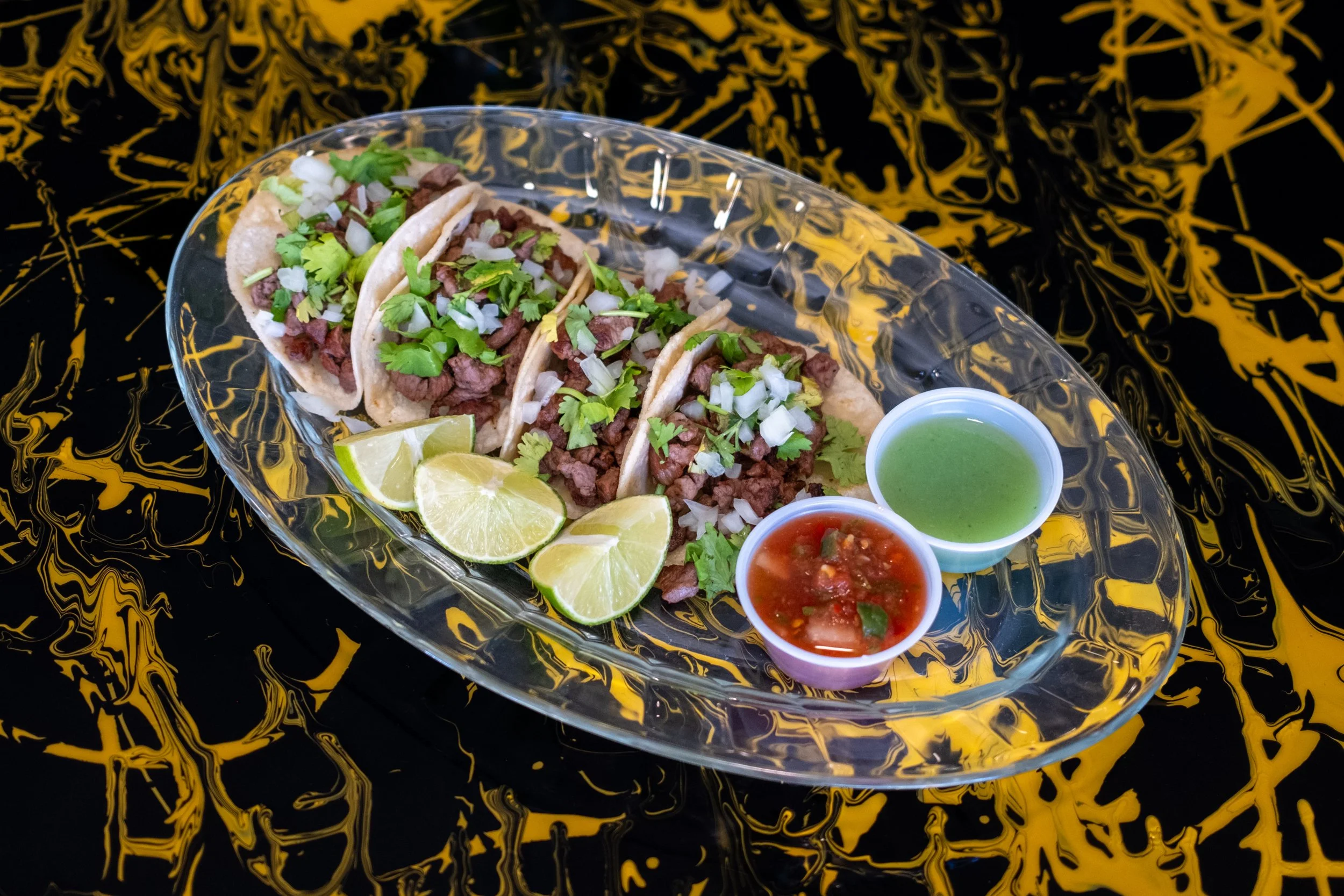 Asada Tacos with cilantro, onions, and lime wedges on a glass plate, accompanied by green and red salsa cups, on a black and yellow patterned tablecloth.