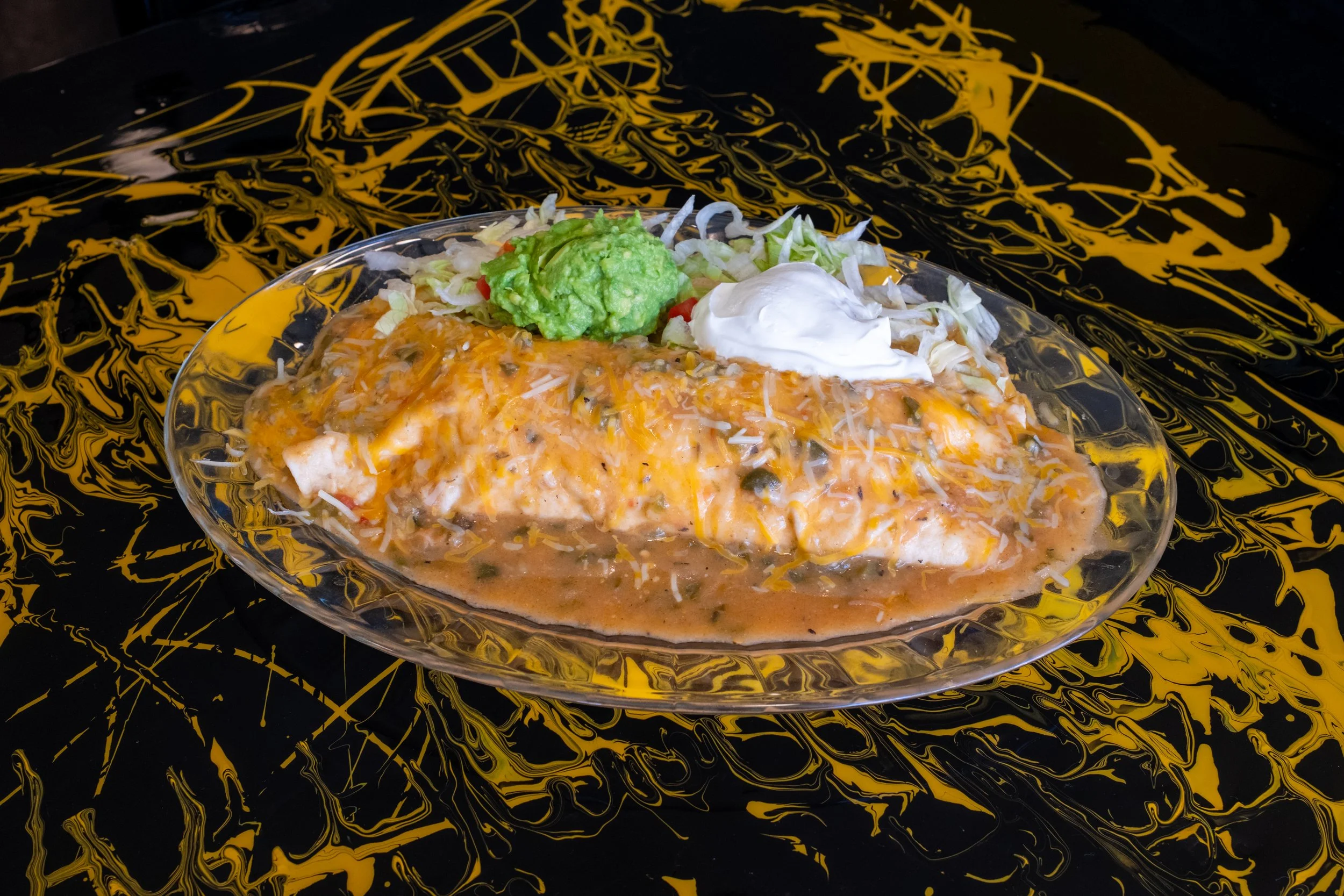 A bowl of Mexican-style food with shredded chicken covered in orange sauce, topped with shredded cheese, guacamole, sour cream, and shredded lettuce, on a black and yellow abstract patterned surface.