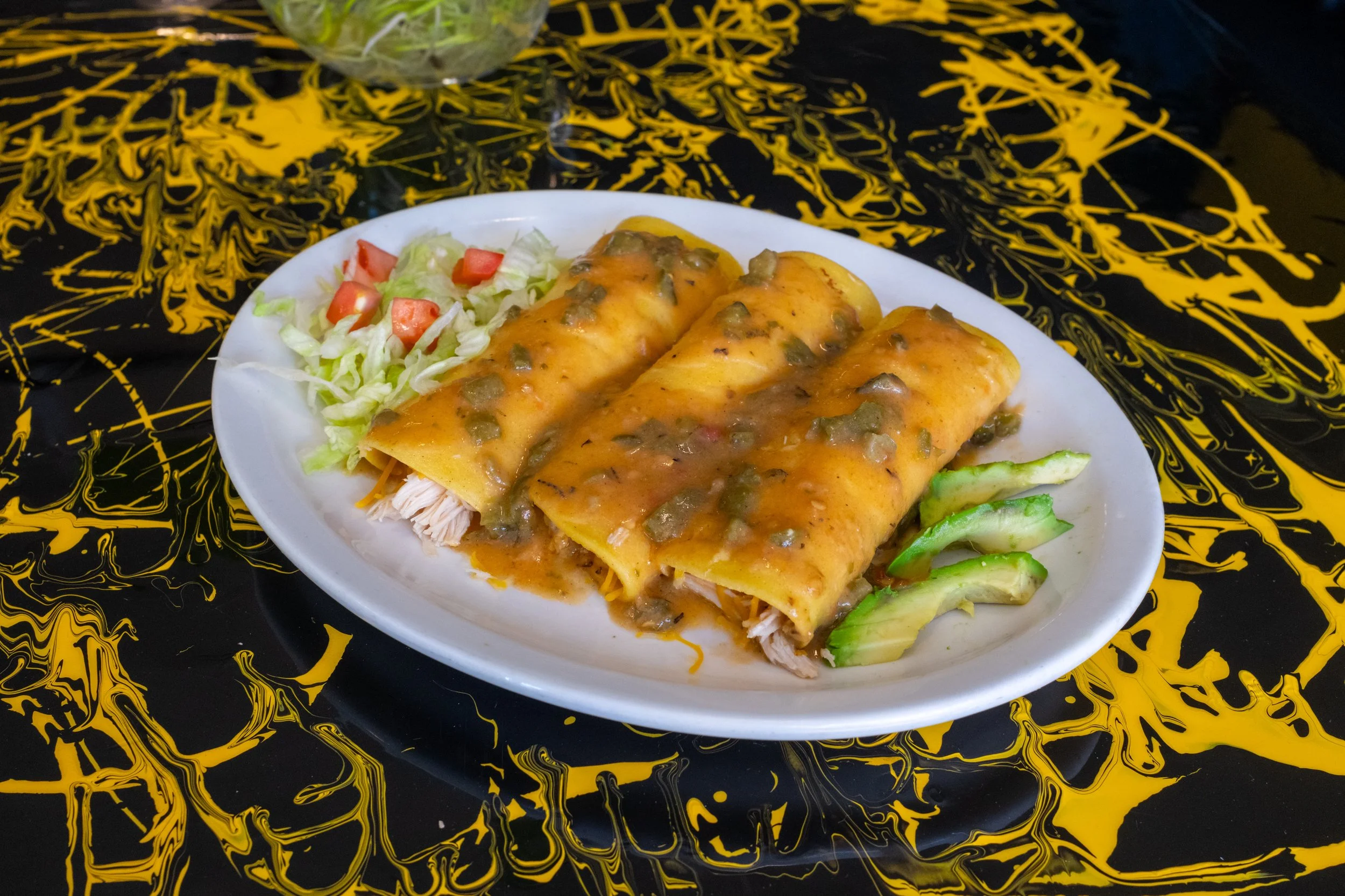 Plate of Mexican food with two beef enchiladas covered in sauce, served with lettuce, tomato, and sliced cucumbers, on a black and yellow abstract tablecloth.