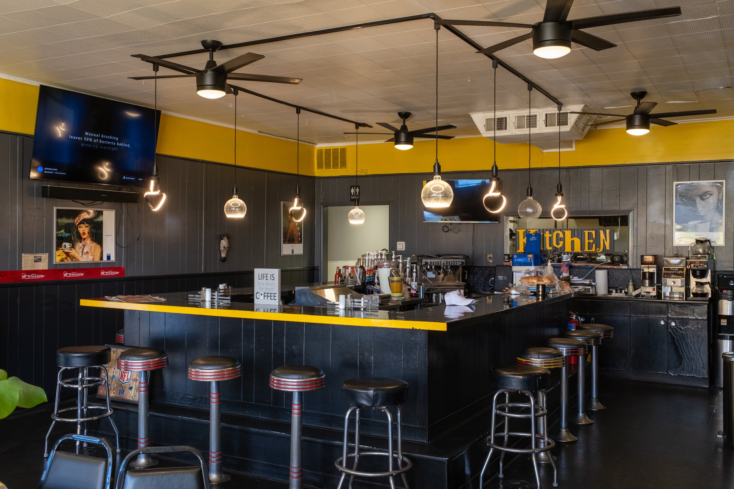 Interior view of a modern diner or cafe with black countertops, barstools, ceiling fans, and hanging light fixtures. There are multiple small appliances and decorations on the counter, and posters on the walls.