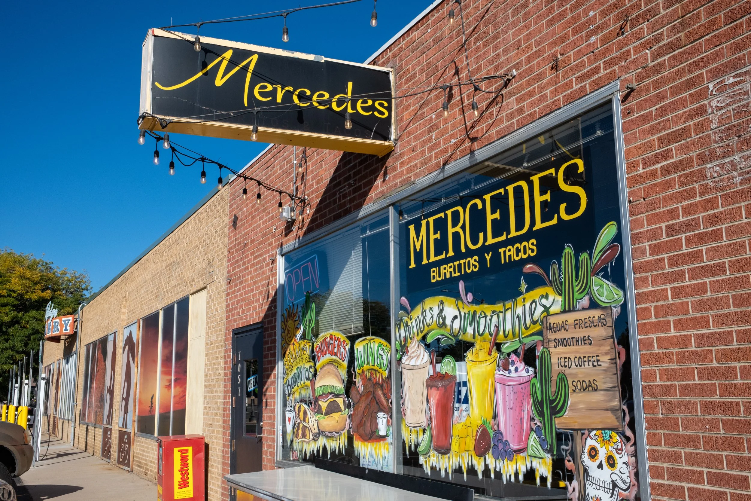 Exterior view of a restaurant named Mercedes, with a colorful menu sign featuring food items like burritos, tacos, burgers, and smoothies, on a brick building under a sunny sky.