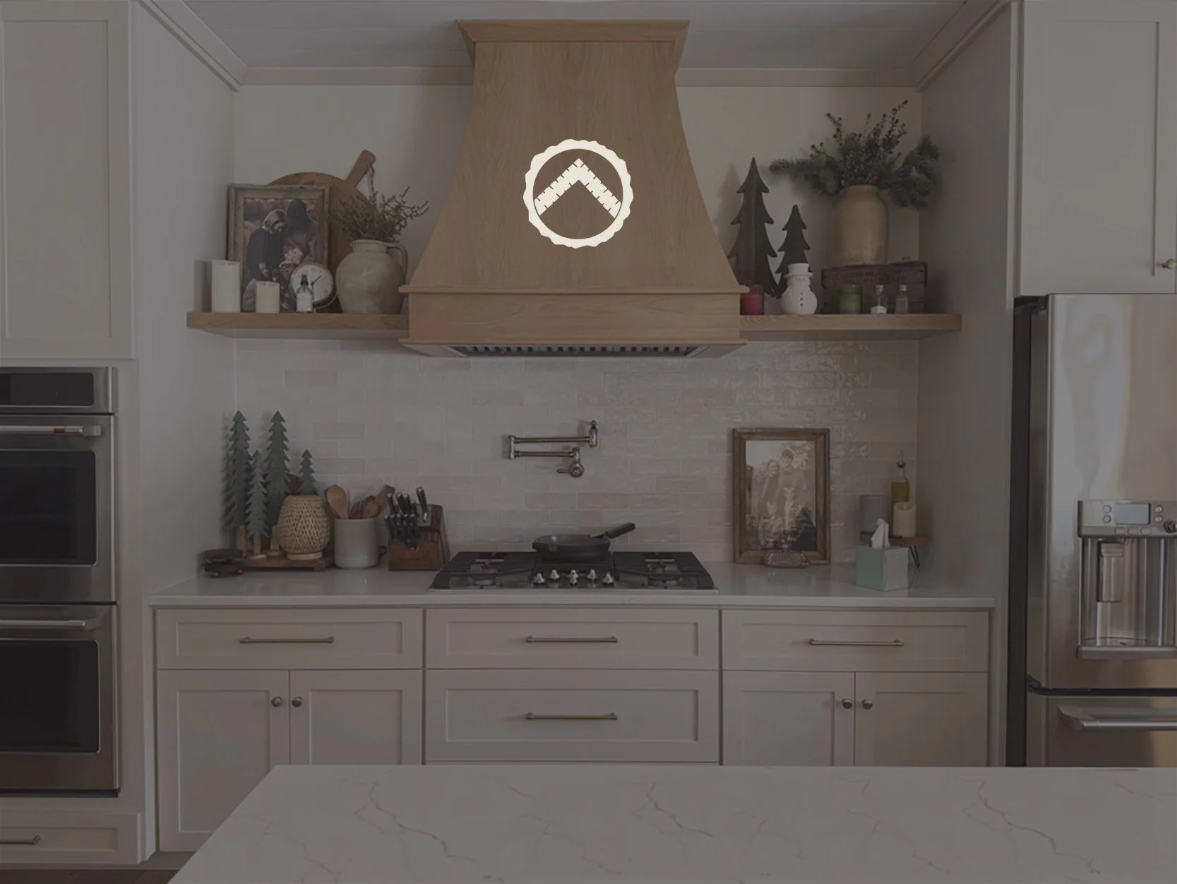 A modern kitchen with white cabinets, a marble countertop, and a gas stove. Decor includes framed photos, candles, small trees, jars, a framed picture, and a soap dispenser. Wooden floating shelves hold vases, greenery, and decorative items. Stainless steel appliances are visible on the left and right sides.