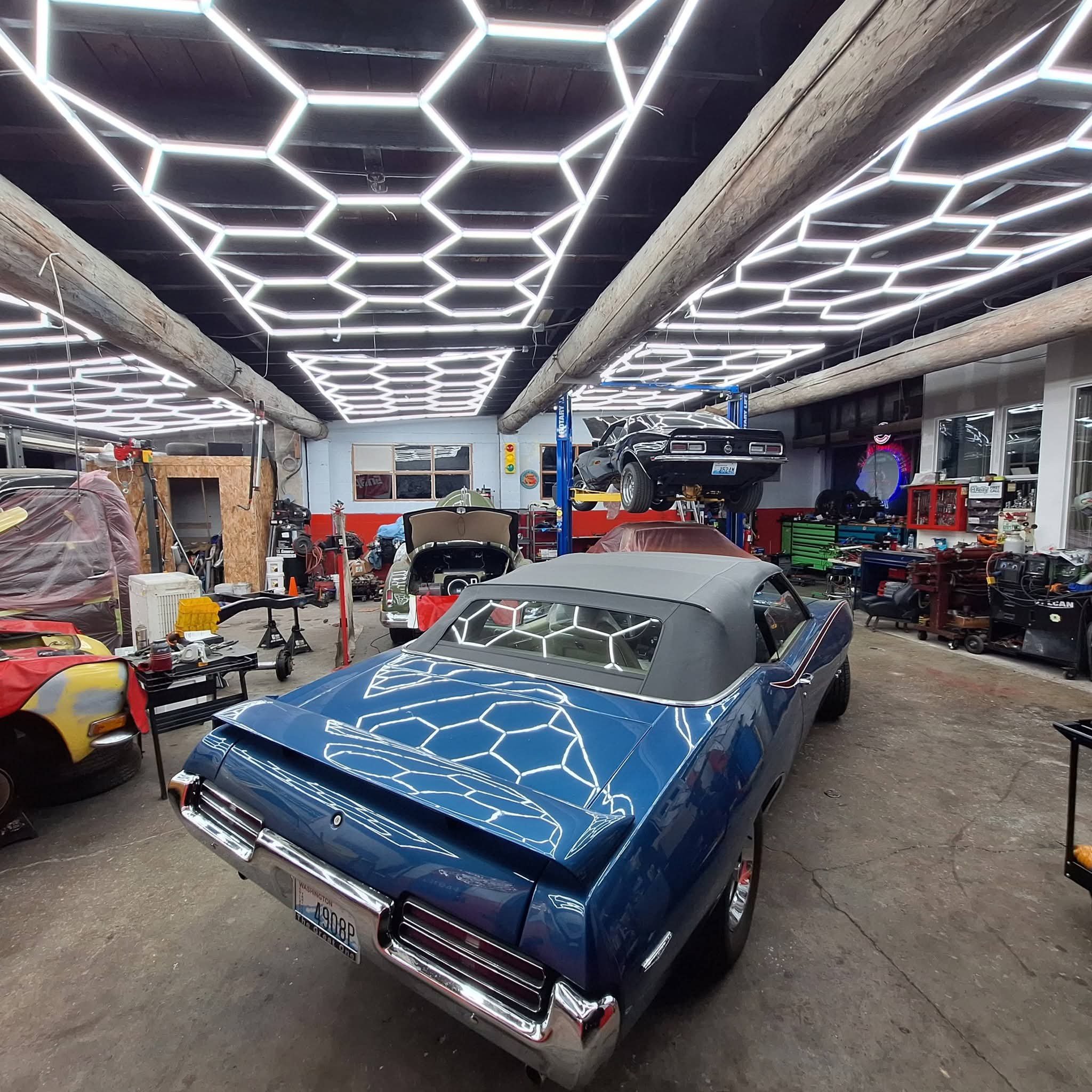 A garage with a retro black muscle car elevated on a lift, surrounded by classic cars and tools. The ceiling has a modern hexagonal LED light pattern, and the garage is filled with various car parts and equipment.
