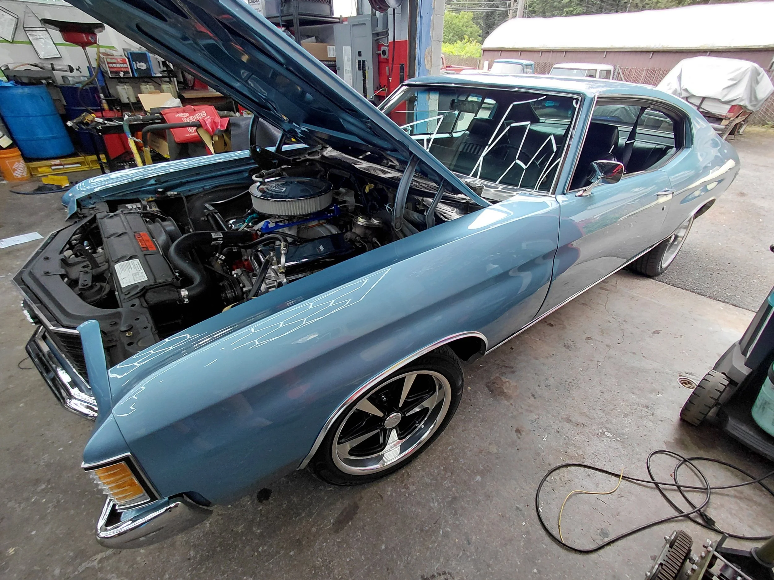 A blue vintage muscle car with its hood open, showing the engine, in a garage or workshop.