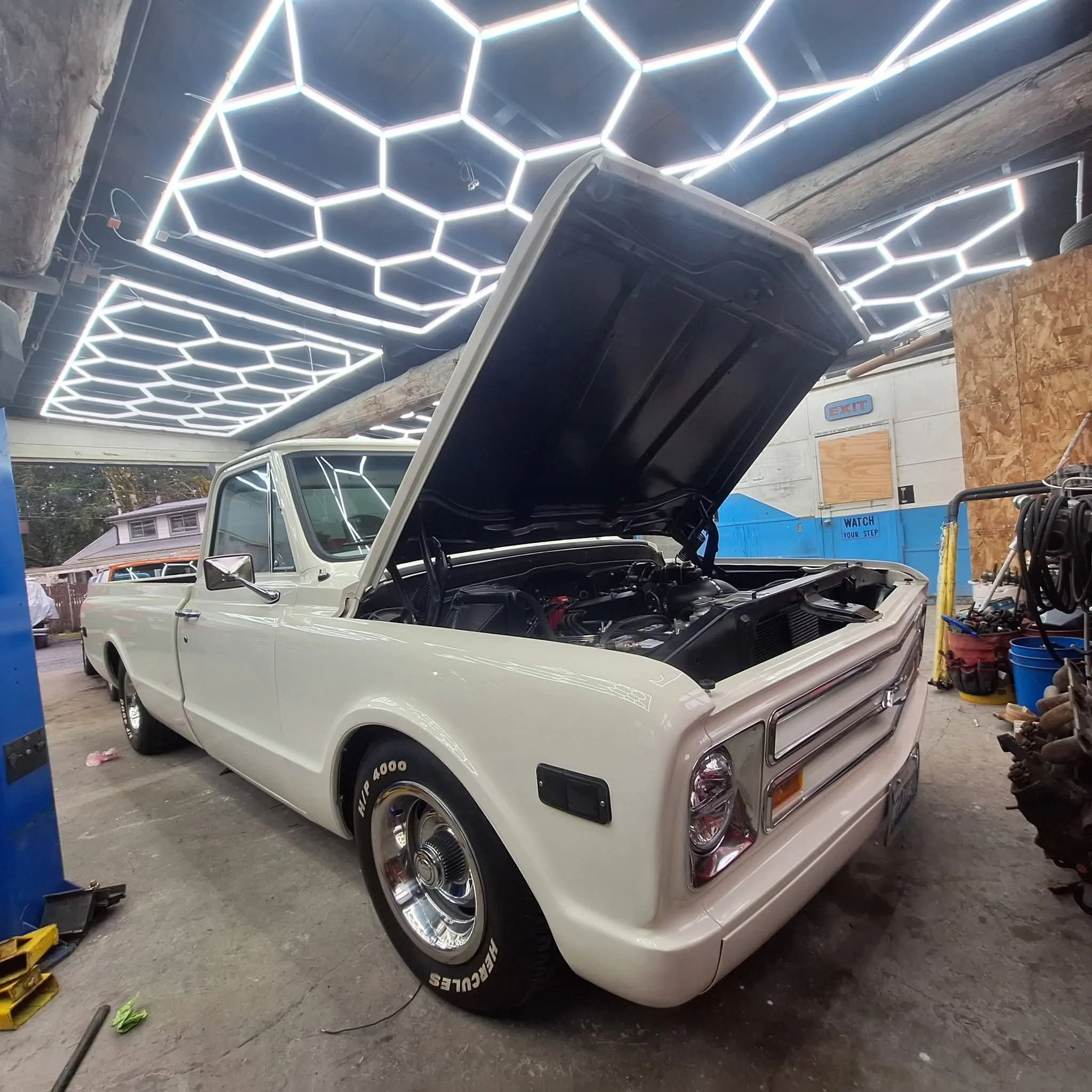 A white vintage car with its hood open inside a garage or workshop with hexagonal LED ceiling lights and various tools and materials around.