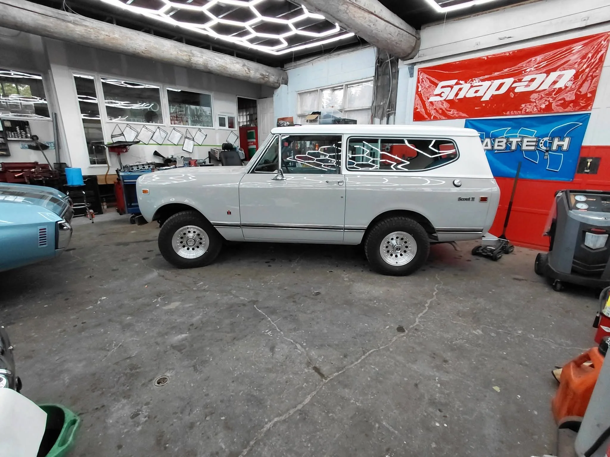 A classic silver compact car is parked inside a garage or workshop space with various tools and banners on the walls, including a large red Snap-On banner.