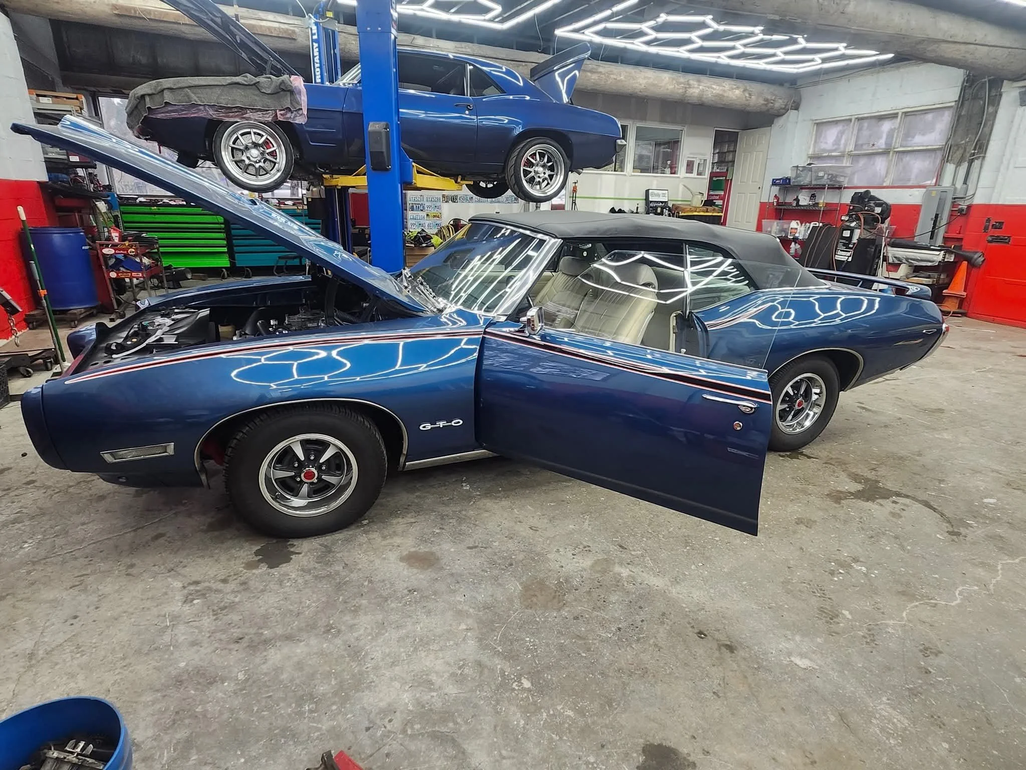 A blue classic car with its hood open and a blue convertible car parked in a garage with a car lift holding another blue car with its hood open. The garage has tools, storage bins, and workbench, with bright overhead lighting.