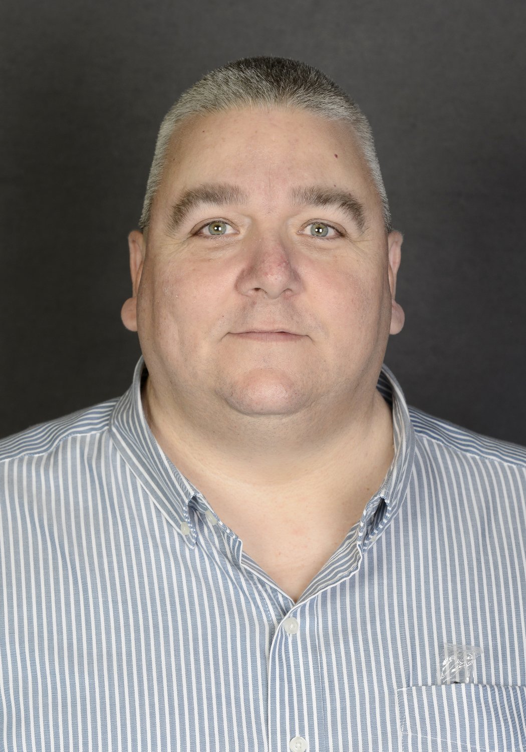 Portrait of a man with short gray hair, light-colored eyes, wearing a striped button-up shirt, against a dark background.