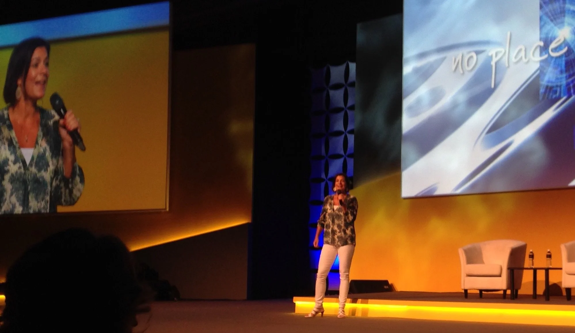 A woman in a floral blazer, white pants, and high heels standing on stage holding a microphone, with a large screen behind her displaying the words 'no place' on a blue background. Another woman in a similar blazer is standing beside her, smiling, with chairs on the stage.