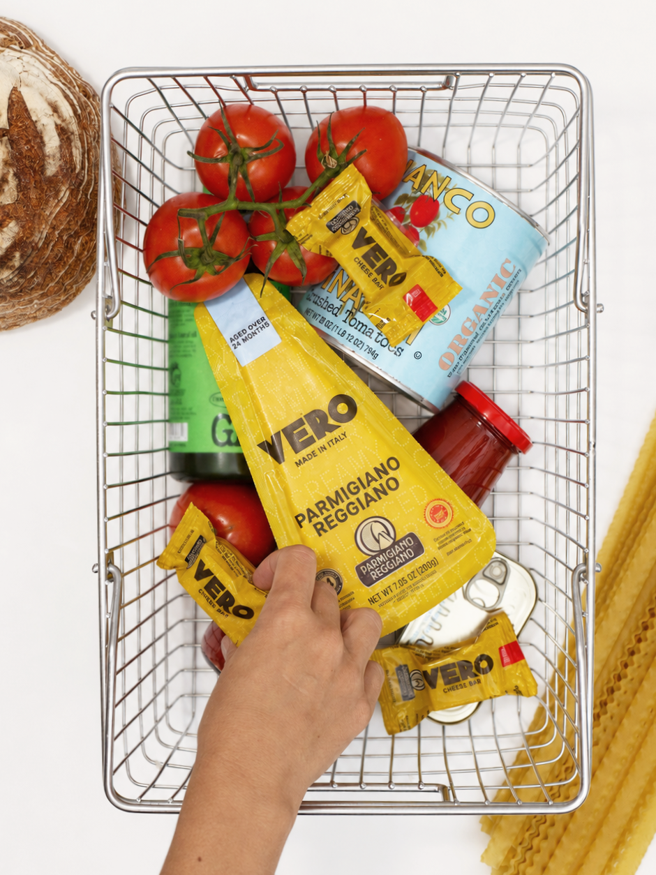 A hand reaching into a metal shopping basket containing fresh tomatoes on the vine, a can of organic crushed tomatoes, various Vero cheese snack bars, a jar of tomato sauce, and a partially visible green can. To the side of the basket is a large piece of cured meat or ham, and on the right side, there are uncooked lasagna noodles.