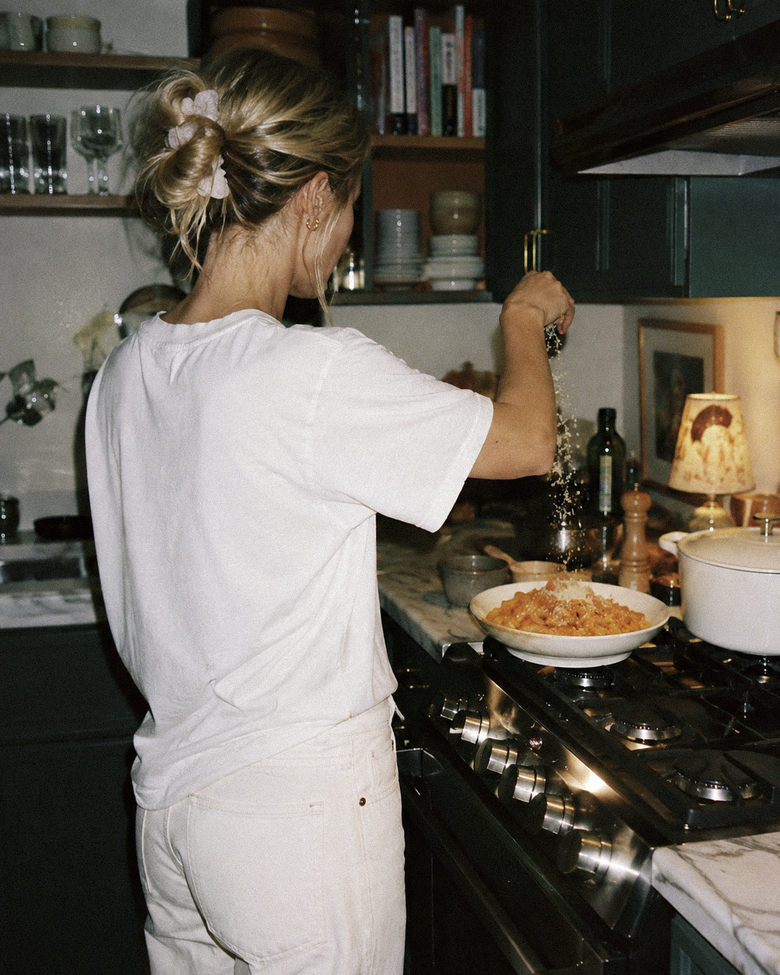A woman with blonde hair in a bun, wearing a white t-shirt and beige pants, seasoning a pasta dish in a kitchen.