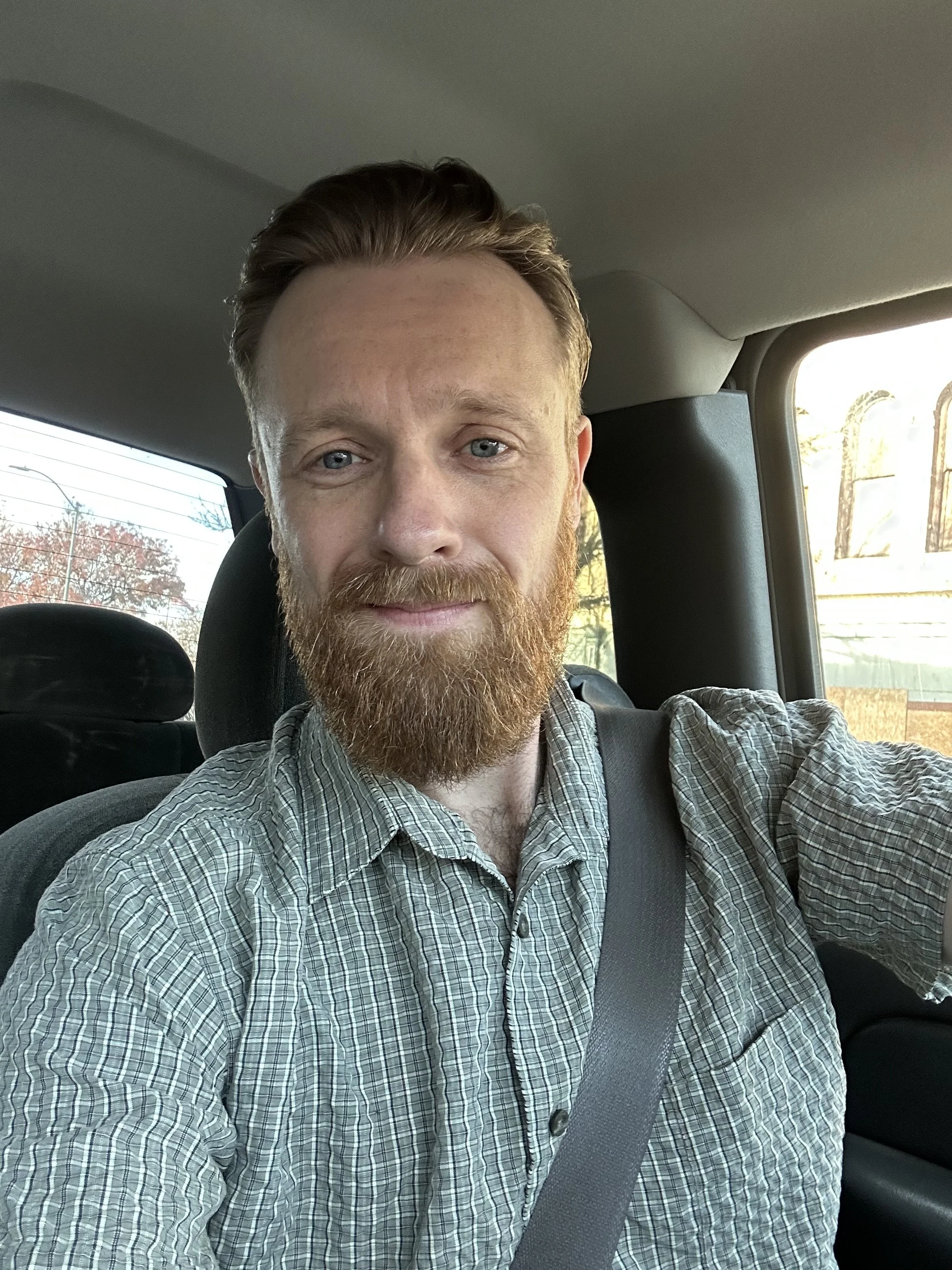 A man with red hair and a beard taking a selfie inside a car while wearing a seatbelt. Background shows a partially cloudy sky and some trees.
