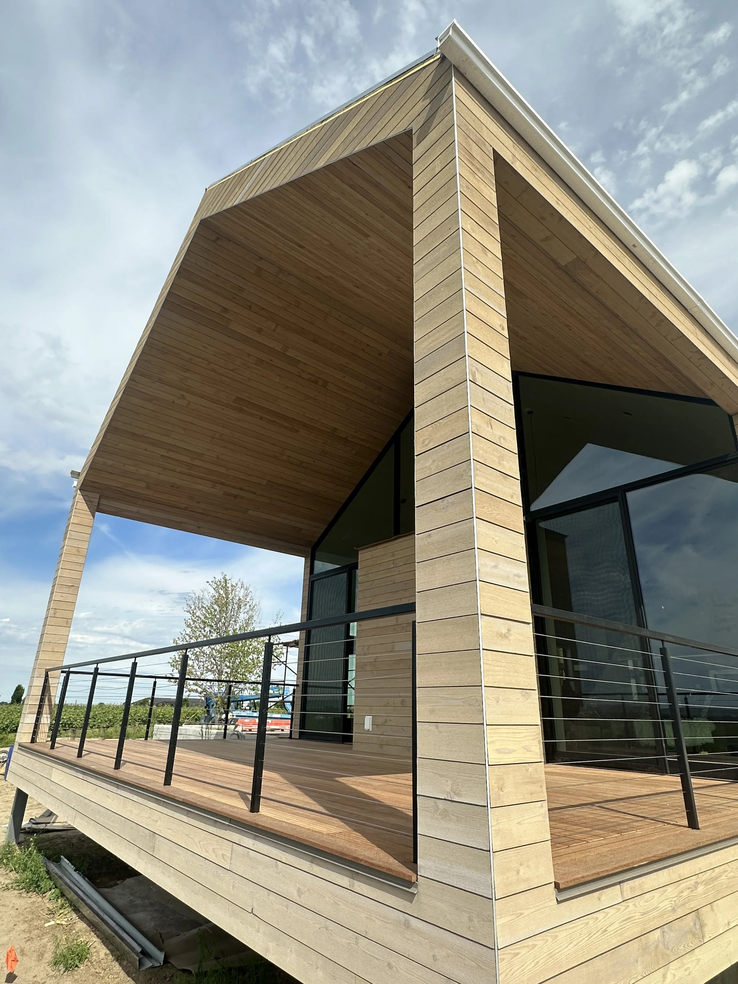 A modern two-story house with a wood exterior, large windows, and a balcony with metal railing, set against a cloudy sky and a rural landscape in the background.